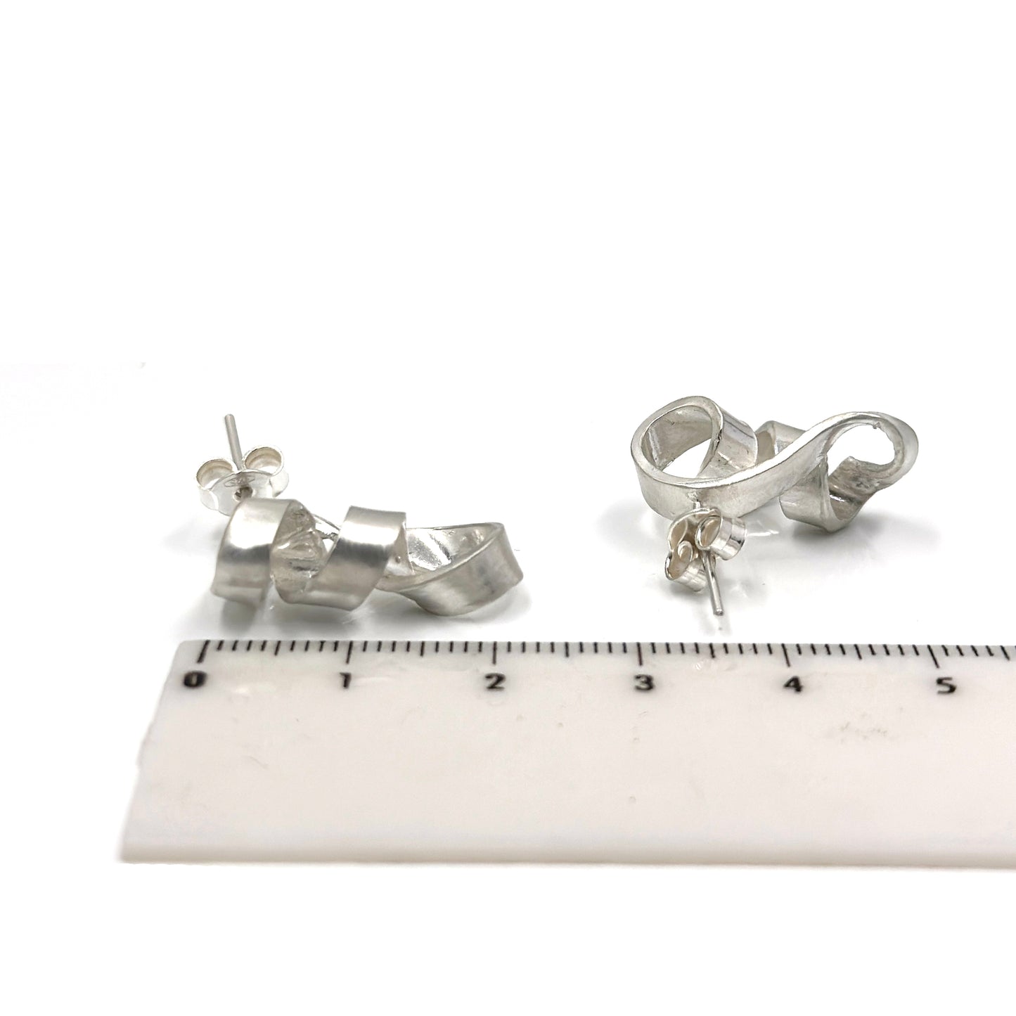 The Archi Small Sterling Silver Earrings by FR | Art Jewellery exhibit an abstract, twisted design. Every earring has a distinctive curved shape reminiscent of a fluid loop with a ribbon-like appearance, highlighting the craftsmanship of handmade jewellery.