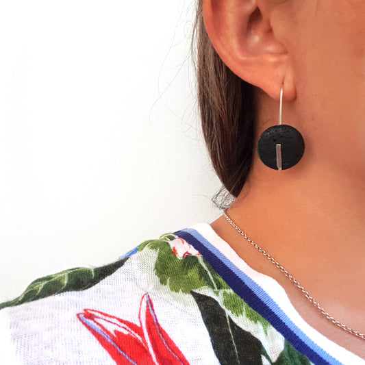 Introducing the Black.o Lava Sterling Silver Earrings by FR | Art Jewellery: These circular earrings showcase a striking textured design fashioned from dark purple volcanic stone, elegantly accented with a sterling silver bar running across the center. The earrings hang gracefully from simple hooks, making them an eye-catching statement piece.