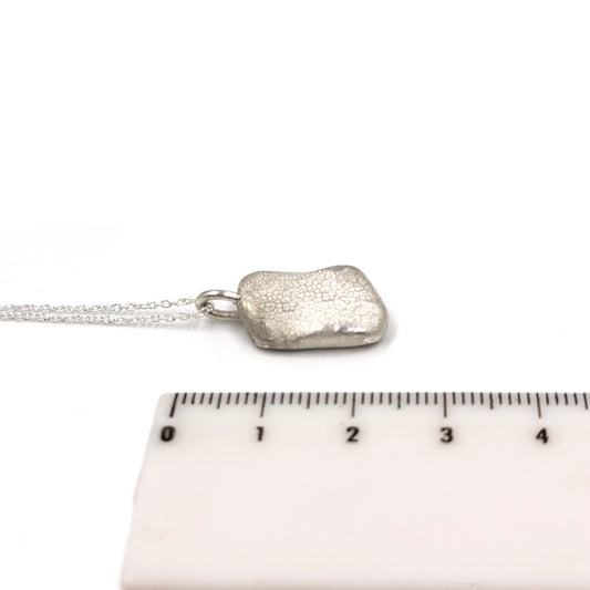 A petite, rectangular silver pendant named "Dog Imprint" by FR | Art Jewellery features a slightly irregular and textured surface. This custom keepsake pendant has a flat loop at the top for easy attachment to a chain or necklace, elegantly set against a plain white background.