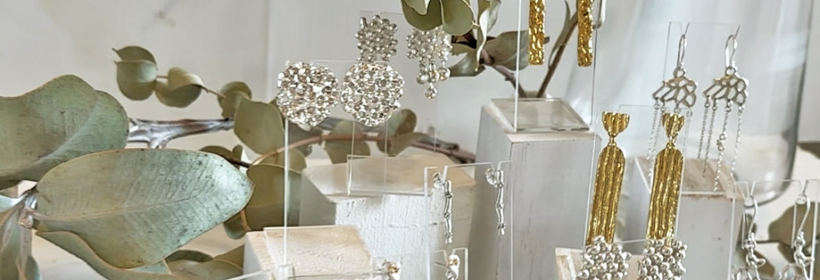 A display of intricate silver and gold earrings arranged on clear and white stands, with large green leaves as backdrop. The earrings include various designs, such as tassels, clusters, and dangling patterns.