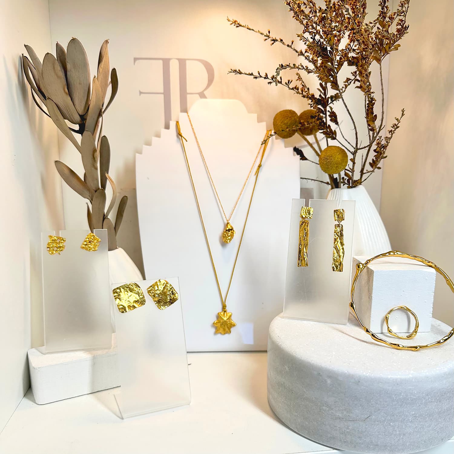 A display of gold jewelry including drop earrings, a geometric necklace, a star-shaped pendant necklace, a bangle bracelet, and a circular ring, arranged on white stands with dried floral décor in the background.