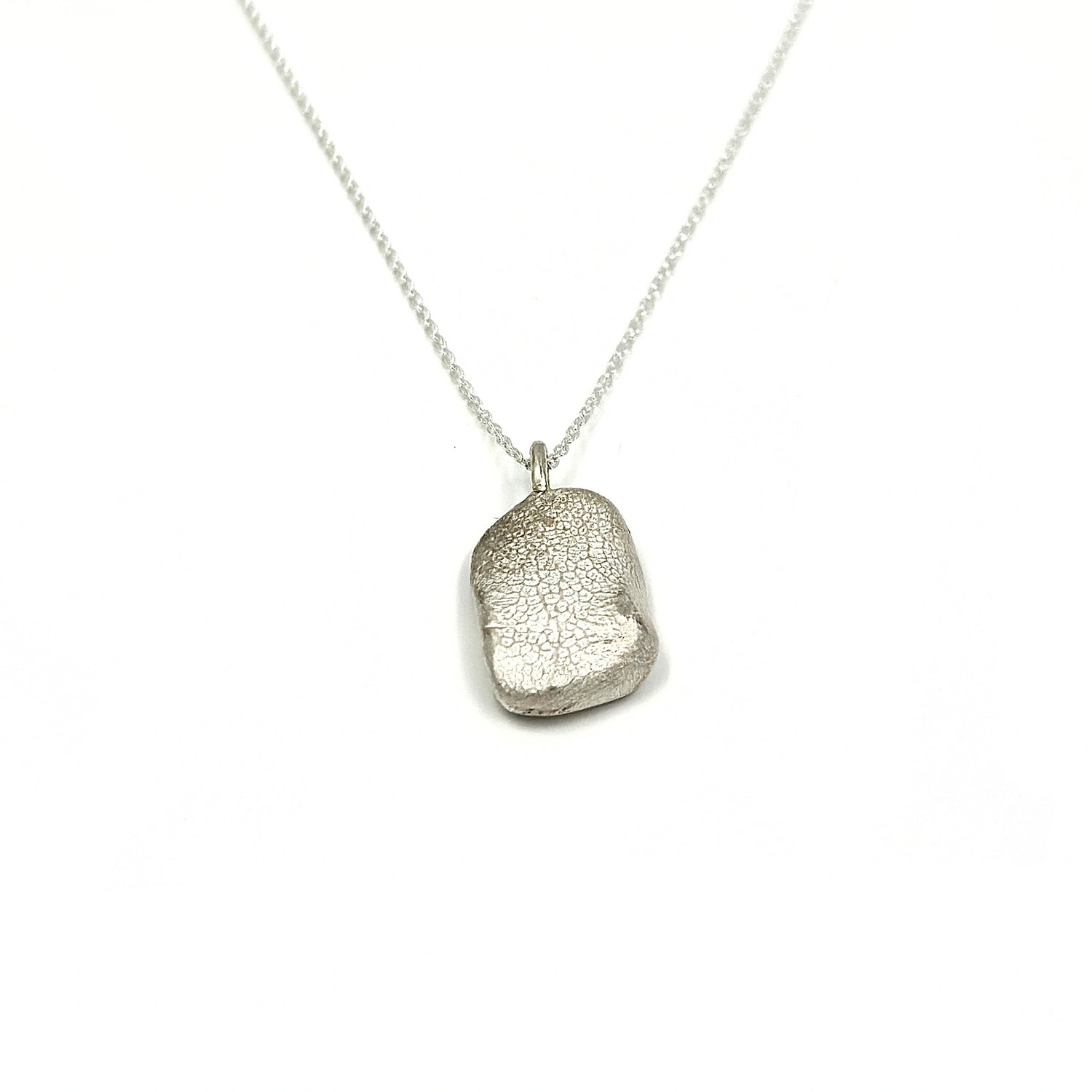 A silver necklace features a delicate chain and a pendant with a textured, asymmetrical rounded shape. The pendant has a subtle, intricate surface design that adds an elegant touch to the minimalist jewelry piece.