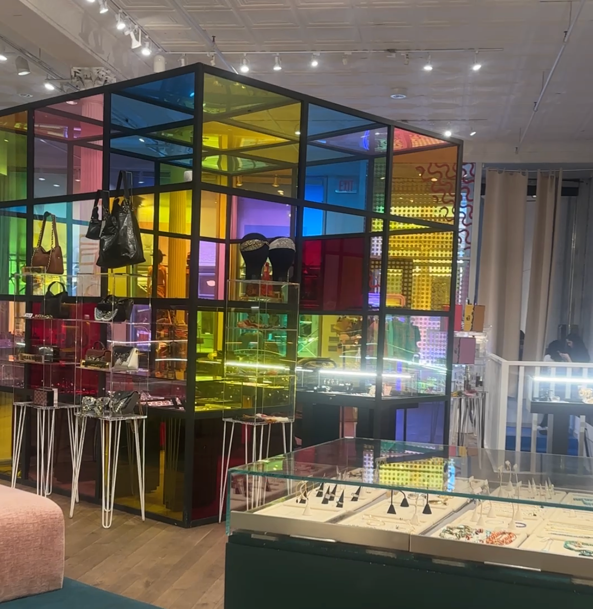 A vibrant retail display features a glass structure with colorful panels, housing handbags and accessories. Jewelry is showcased in glass cases nearby, under bright ceiling lights.