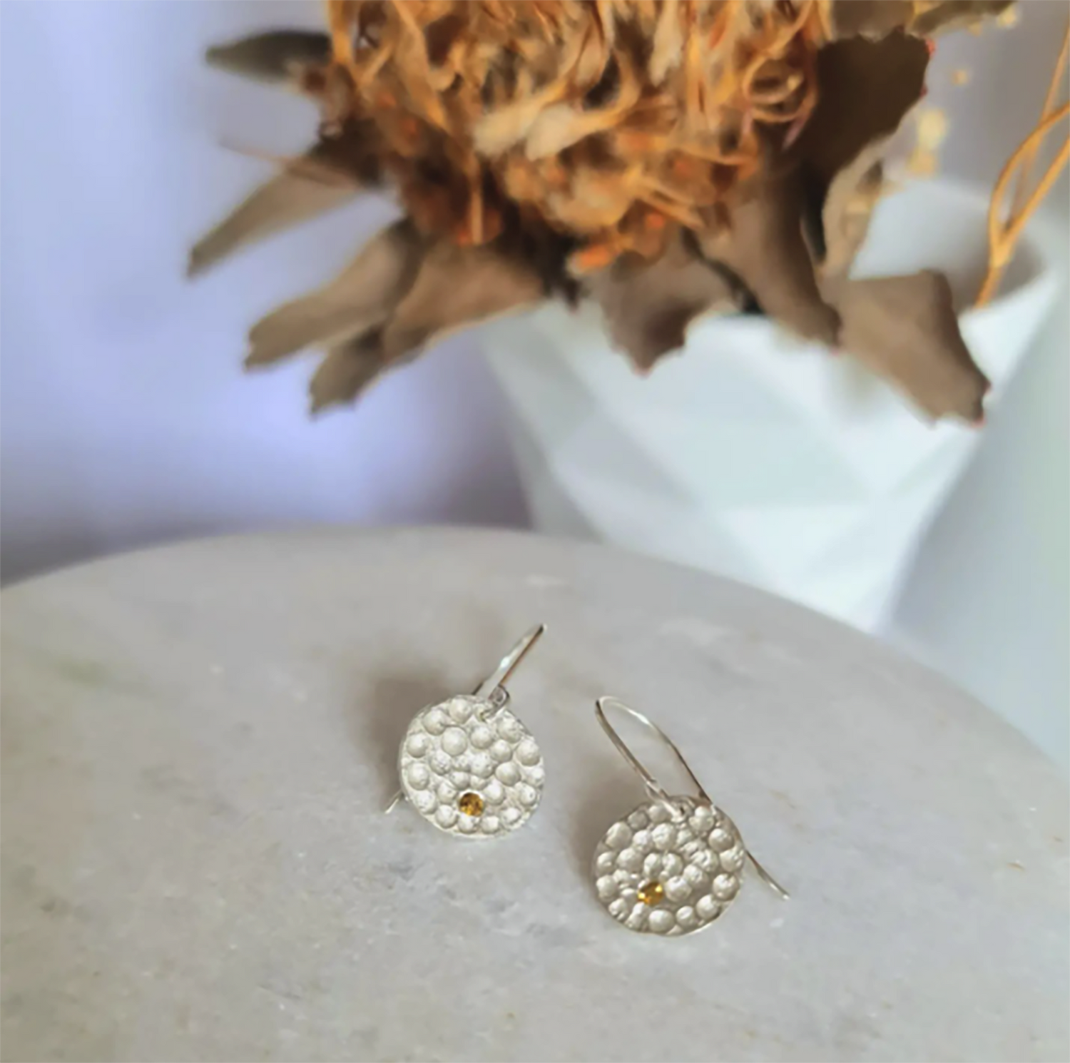 Close-up of textured silver earrings with small gold accents on a smooth, round surface. A blurred dried flower arrangement in a white vase is in the background.