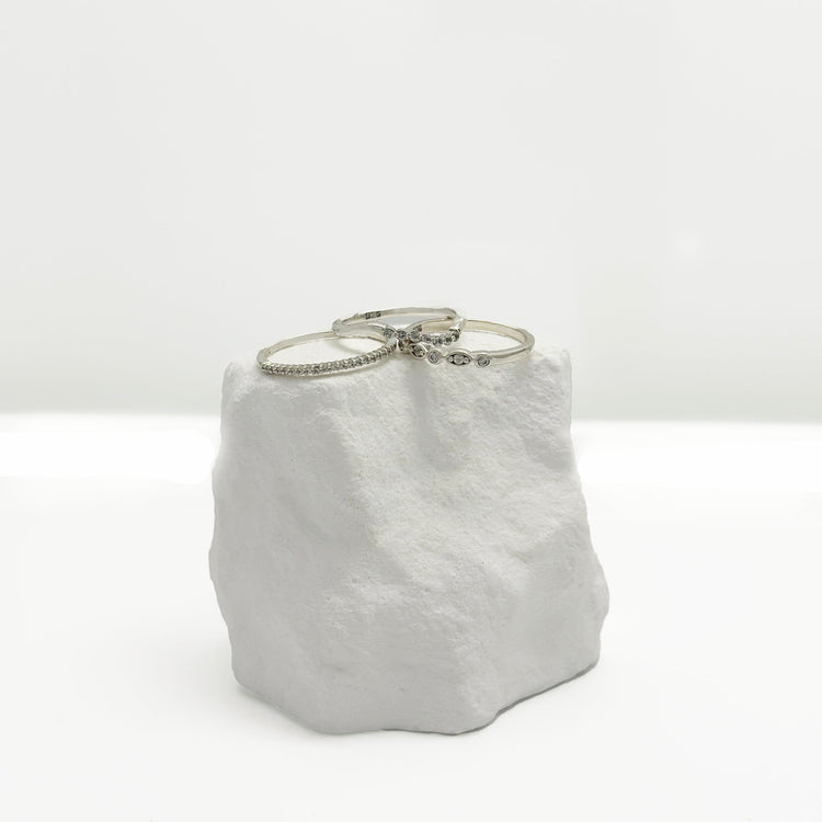 Two silver bracelets, one with small stones and the other with a delicate leaf design, displayed on a textured white stone against a plain white background.