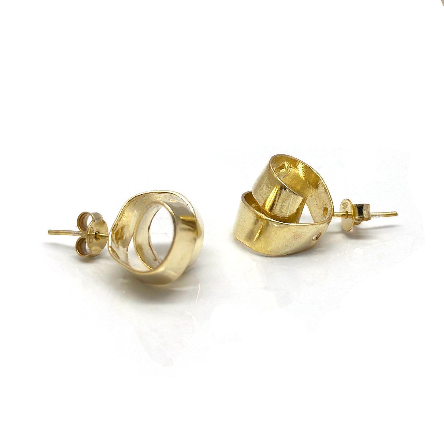 A pair of FR | Art Jewellery's Mini Archi Studs, featuring a handmade, gold-plated, slightly curved ribbon-like design set against a white background. The studs boast a glossy finish that beautifully reflects light.