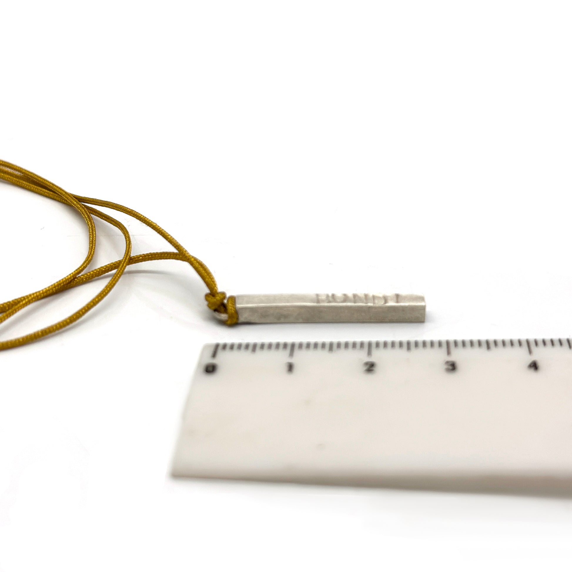 The Bondi Necklace from FR | Art Jewellery features a thin, rectangular metallic pendant with engraved text, hanging from a mustard-yellow cord. This piece of handmade jewellery boasts a smooth, slightly shiny surface and is elegantly suspended against a white background.