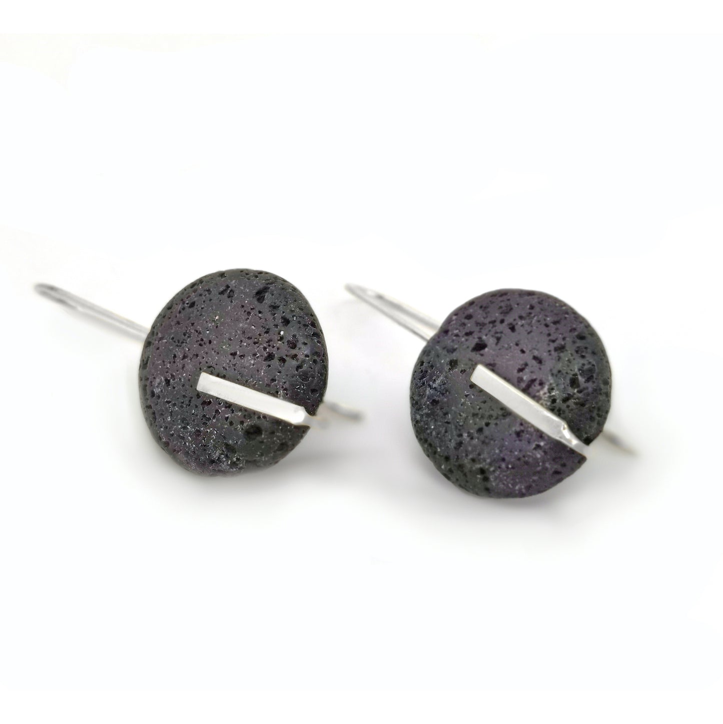 The Black.o Lava Earrings by FR | Art Jewellery feature round, dark gray, porous volcanic stones that resemble lava rock. These distinctive stones are adorned with a thin, shiny metallic bar running horizontally across the center. Crafted with sterling silver hooks, these earrings offer an elegant touch and ensure easy wearing.