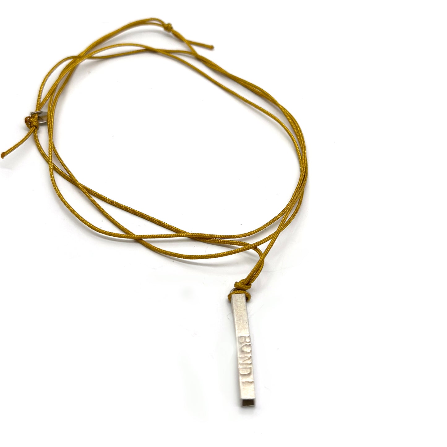 The Bondi Necklace from FR | Art Jewellery features a thin, rectangular metallic pendant with engraved text, hanging from a mustard-yellow cord. This piece of handmade jewellery boasts a smooth, slightly shiny surface and is elegantly suspended against a white background.