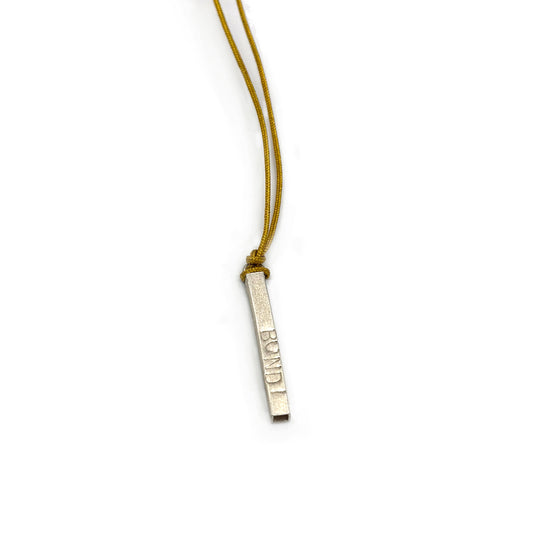 The Bondi Necklace from FR | Art Jewellery features a thin, rectangular metallic pendant with engraved text, hanging from a mustard-yellow cord. This piece of handmade jewellery boasts a smooth, slightly shiny surface and is elegantly suspended against a white background.