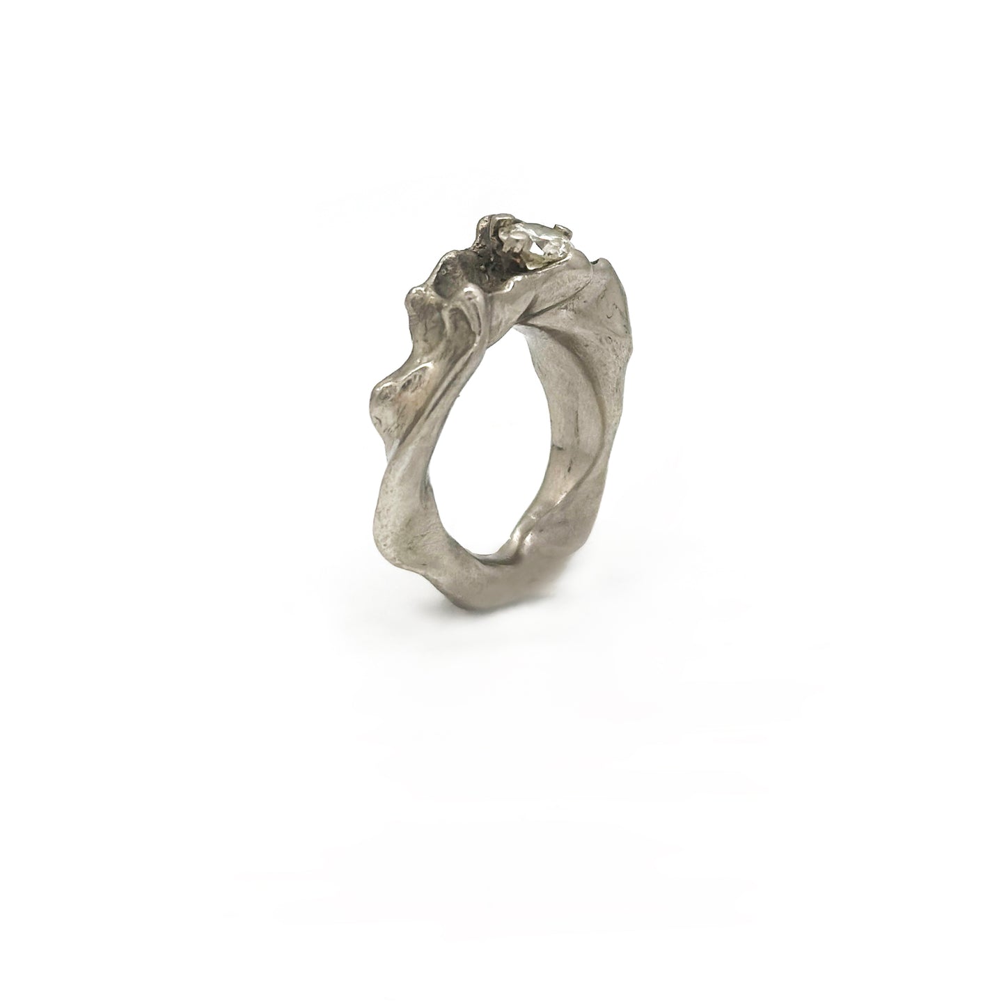 Displayed against a plain white background is the Caracoles Diamond Ring from FR | Art Jewellery's handmade collection. This silver-colored ring features a uniquely textured, wavy band with a small, sparkling diamond set on top. Its organic, fluid design evokes the impression of natural formations.