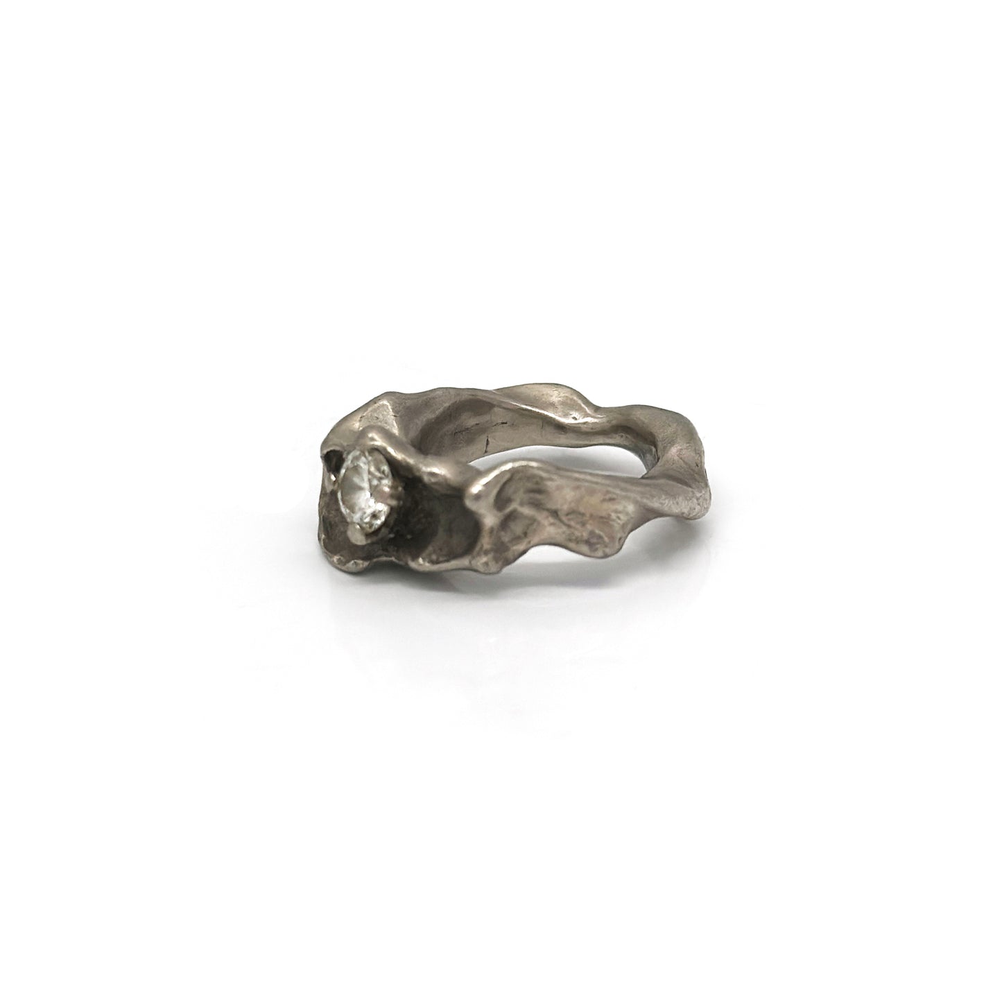 Displayed against a plain white background is the Caracoles Diamond Ring from FR | Art Jewellery's handmade collection. This silver-colored ring features a uniquely textured, wavy band with a small, sparkling diamond set on top. Its organic, fluid design evokes the impression of natural formations.