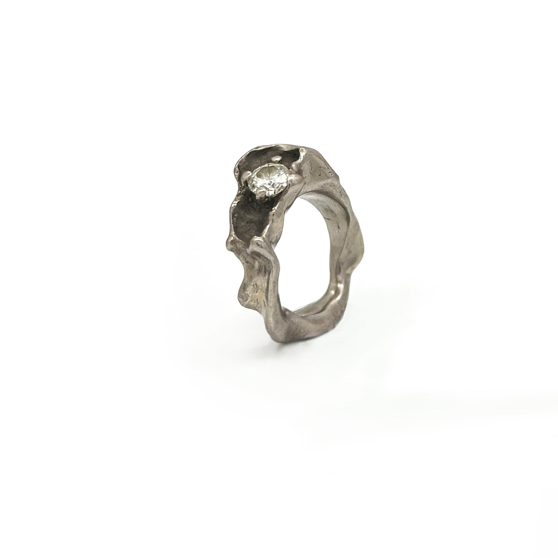 Displayed against a plain white background is the Caracoles Diamond Ring from FR | Art Jewellery's handmade collection. This silver-colored ring features a uniquely textured, wavy band with a small, sparkling diamond set on top. Its organic, fluid design evokes the impression of natural formations.