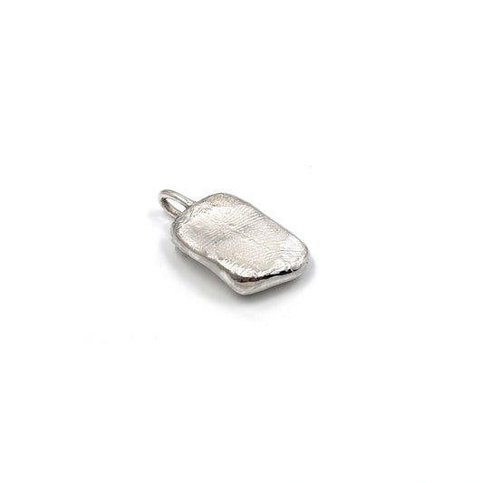 A petite, rectangular silver pendant named "Dog Imprint" by FR | Art Jewellery features a slightly irregular and textured surface. This custom keepsake pendant has a flat loop at the top for easy attachment to a chain or necklace, elegantly set against a plain white background.