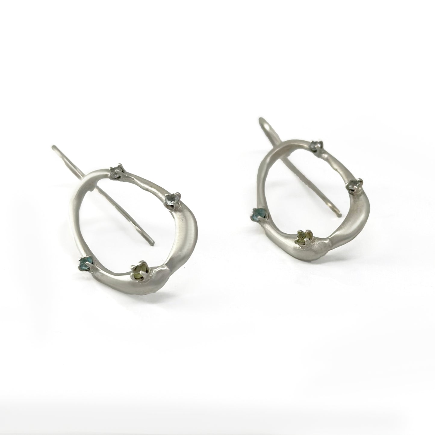 Here is the revised sentence based on the given product data:

A pair of sterling silver Drops Earrings with an irregular open hoop design, each adorned with small green gemstones and tiny star-shaped accents. Part of the Blossom series from the Inspired by Nature Collection by FR | Art Jewellery, these earrings have a hook closure and are displayed against a plain white background.