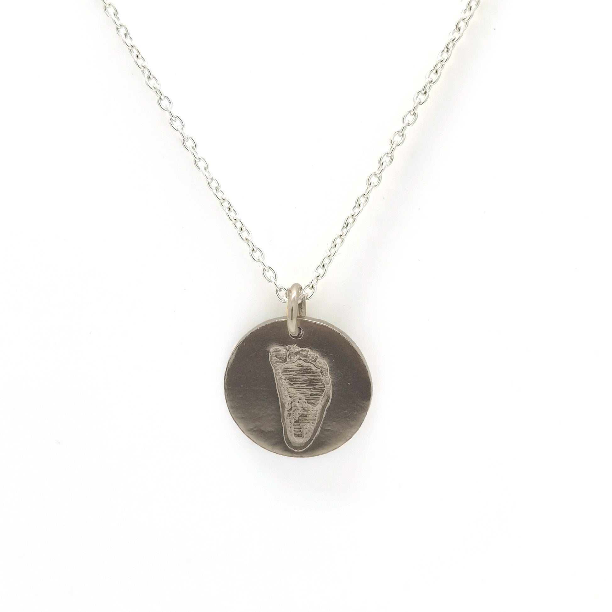 Introducing the Footprint Necklace from FR | Art Jewellery: part of our exquisite Footprint Collection, this silver necklace boasts a simple chain adorned with a circular baby footprint pendant. The pendant features an engraved imprint of a small footprint, offering a personal and delicate touch to this beautiful piece. With its plain white background, this personalized keepsake stands out as a perfect memento.