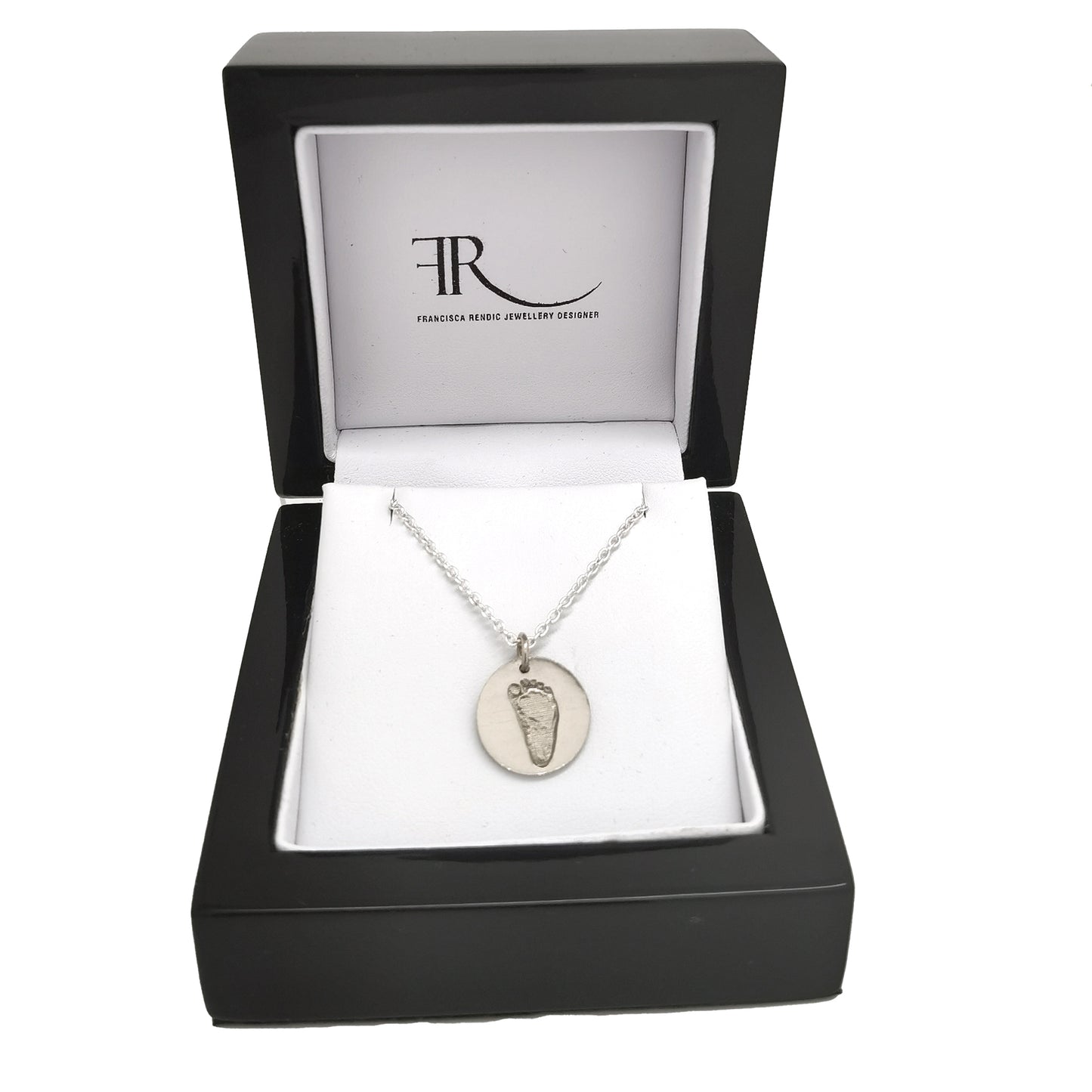 Introducing the Footprint Necklace from FR | Art Jewellery: part of our exquisite Footprint Collection, this silver necklace boasts a simple chain adorned with a circular baby footprint pendant. The pendant features an engraved imprint of a small footprint, offering a personal and delicate touch to this beautiful piece. With its plain white background, this personalized keepsake stands out as a perfect memento.