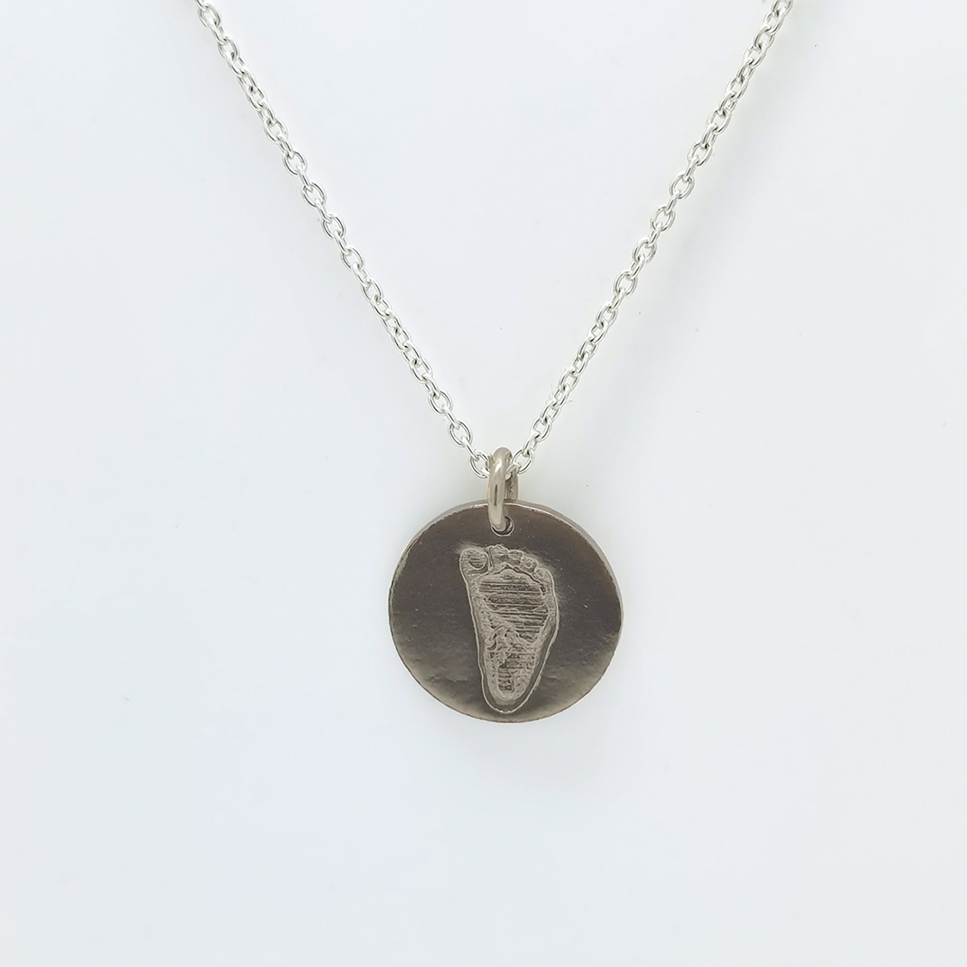 Introducing the Footprint Necklace from FR | Art Jewellery: part of our exquisite Footprint Collection, this silver necklace boasts a simple chain adorned with a circular baby footprint pendant. The pendant features an engraved imprint of a small footprint, offering a personal and delicate touch to this beautiful piece. With its plain white background, this personalized keepsake stands out as a perfect memento.