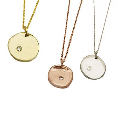 Three round pendant necklaces are displayed against a white background. The pendants are gold, rose gold, and silver, each featuring a small central diamond and matching delicate chains.