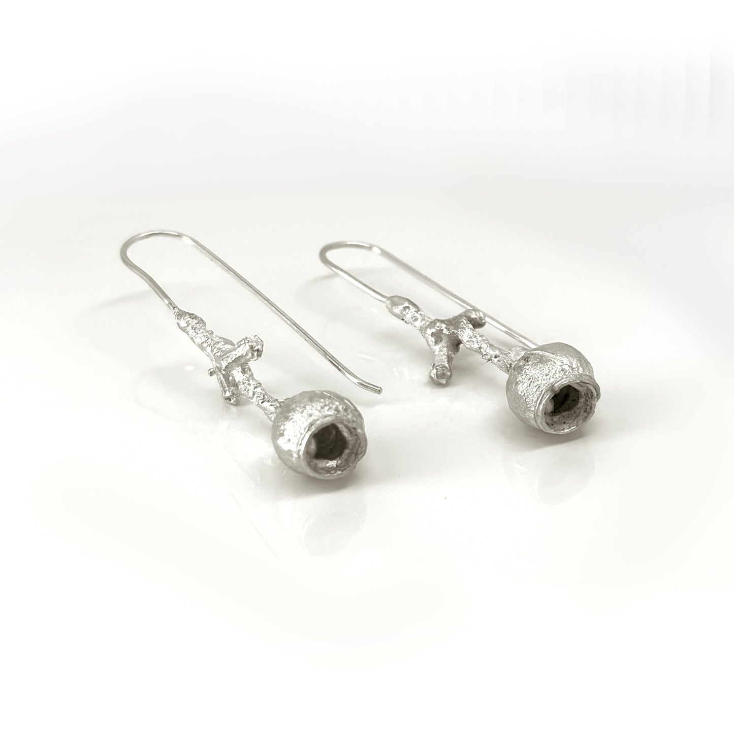 The Gummies Stick Earrings by FR | Art Jewellery are a pair of silver drop earrings with a unique design featuring irregularly shaped hollow structures at the bottom and delicate, textured rods connecting to simple ear hooks. These handmade jewelry pieces are displayed against a clean, white background.