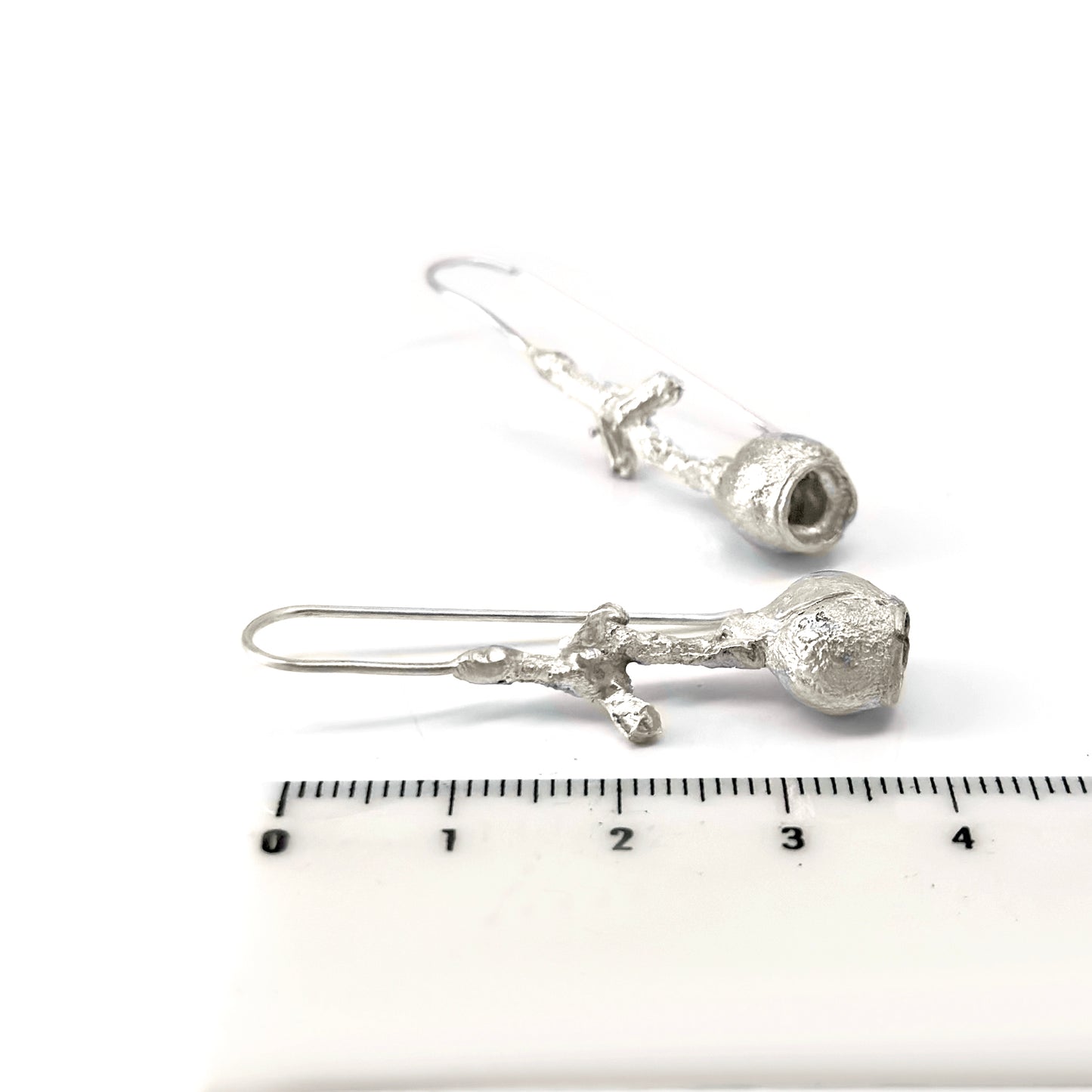 The Gummies Stick Earrings by FR | Art Jewellery are a pair of silver drop earrings with a unique design featuring irregularly shaped hollow structures at the bottom and delicate, textured rods connecting to simple ear hooks. These handmade jewelry pieces are displayed against a clean, white background.