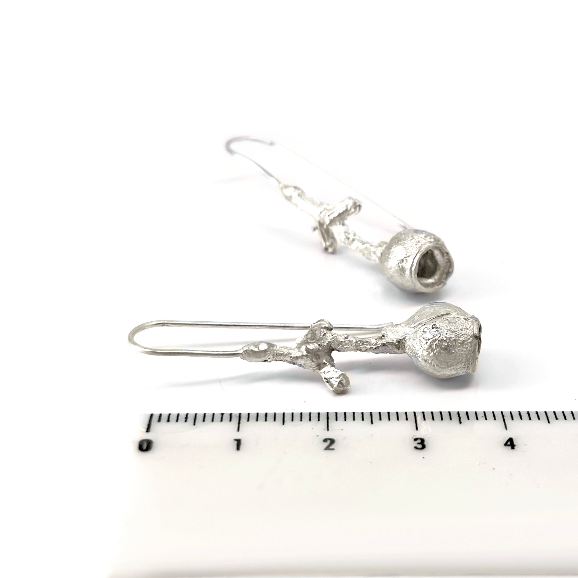 The Gummies Stick Earrings by FR | Art Jewellery are a pair of silver drop earrings with a unique design featuring irregularly shaped hollow structures at the bottom and delicate, textured rods connecting to simple ear hooks. These handmade jewelry pieces are displayed against a clean, white background.