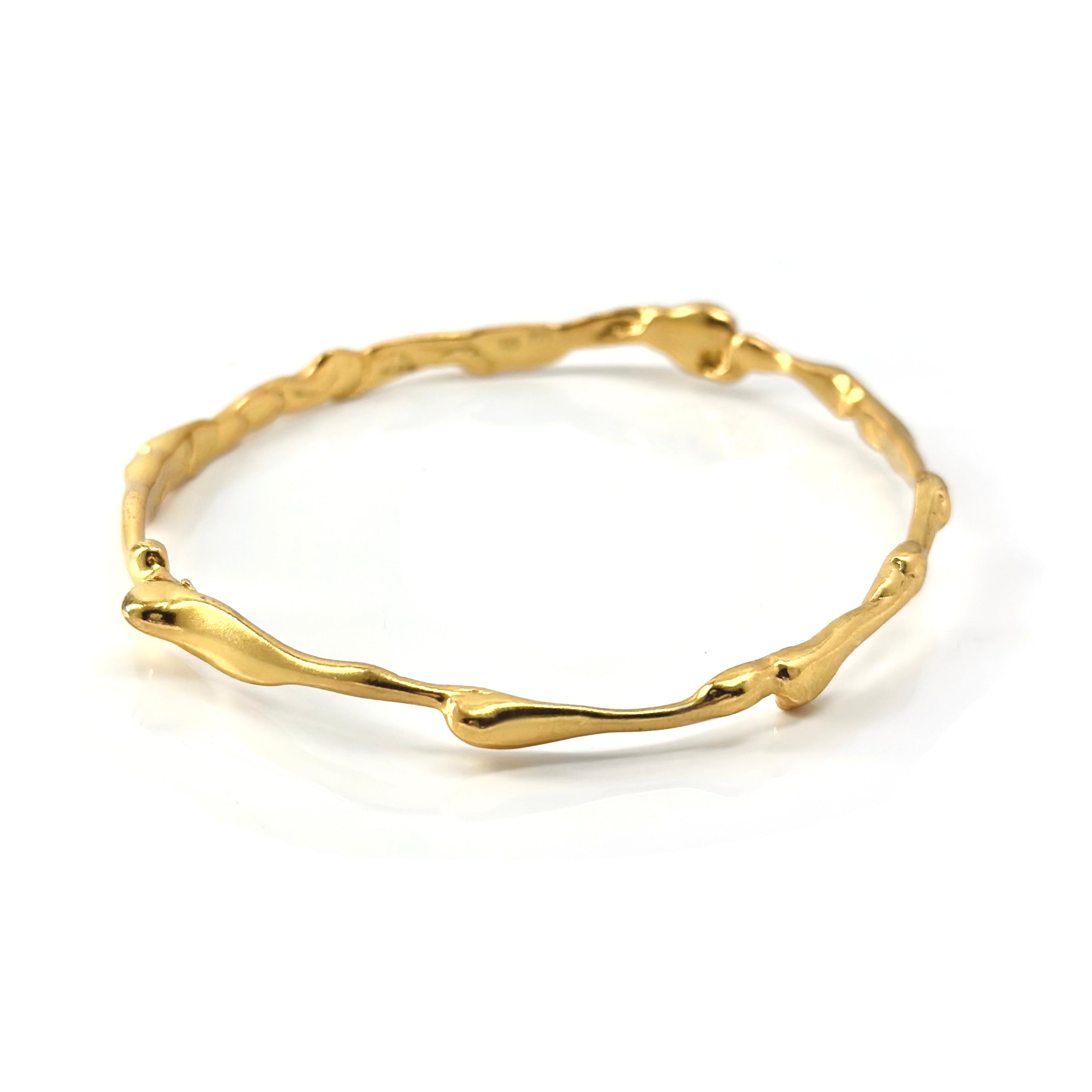 A gold bangle with an organic, irregular design. The bangle features a smooth, wavy texture that mimics the natural contours and imperfections of branches or twigs, creating a modern yet rustic appearance. The gold has a shiny finish, reflecting light elegantly.