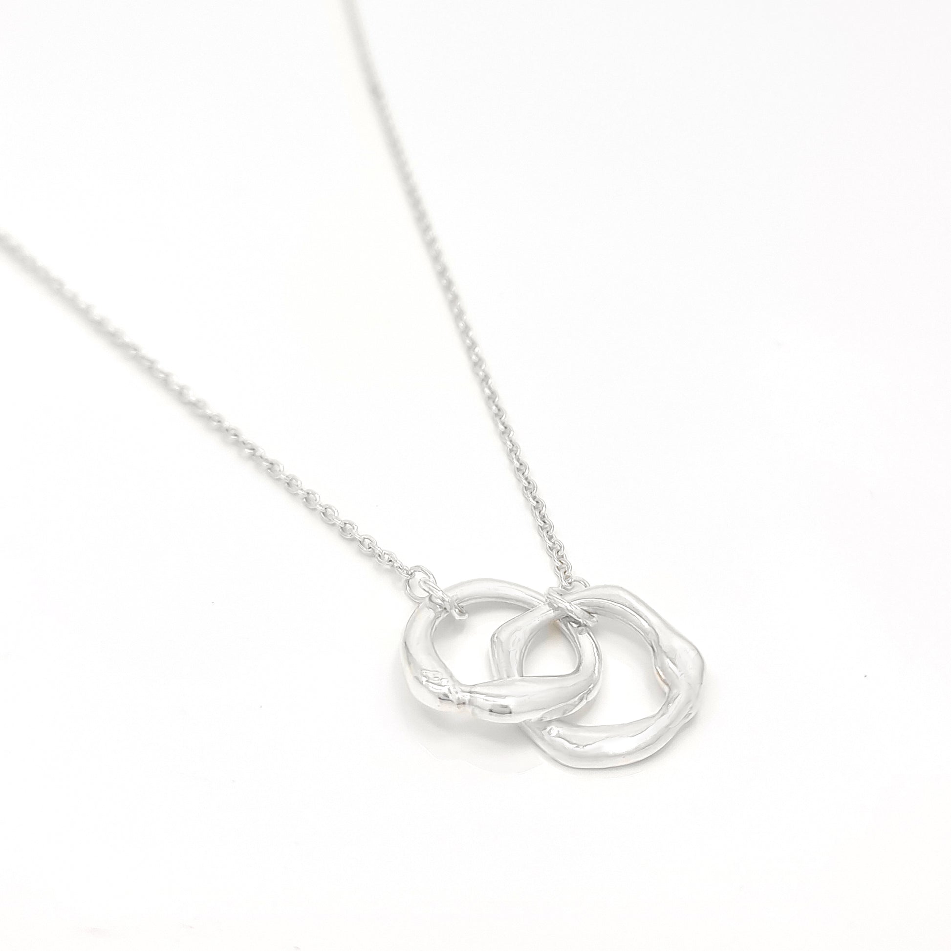 The Hoop Necklace by FR | Art Jewellery is a silver piece with a delicate chain that features two interlocking circular pendants. The pendants have a slightly irregular, textured appearance, creating a modern yet organic look. This handmade jewellery piece is laid flat against a plain white background.