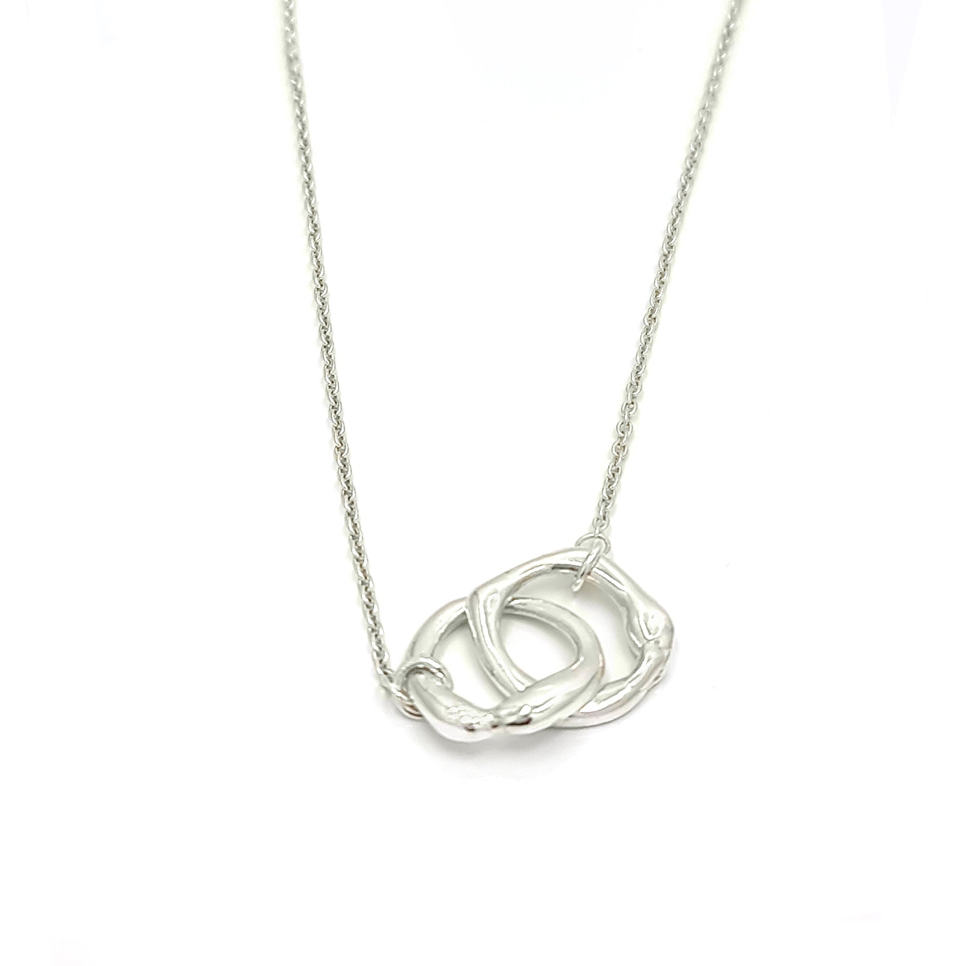 The Hoop Necklace by FR | Art Jewellery is a silver piece with a delicate chain that features two interlocking circular pendants. The pendants have a slightly irregular, textured appearance, creating a modern yet organic look. This handmade jewellery piece is laid flat against a plain white background.