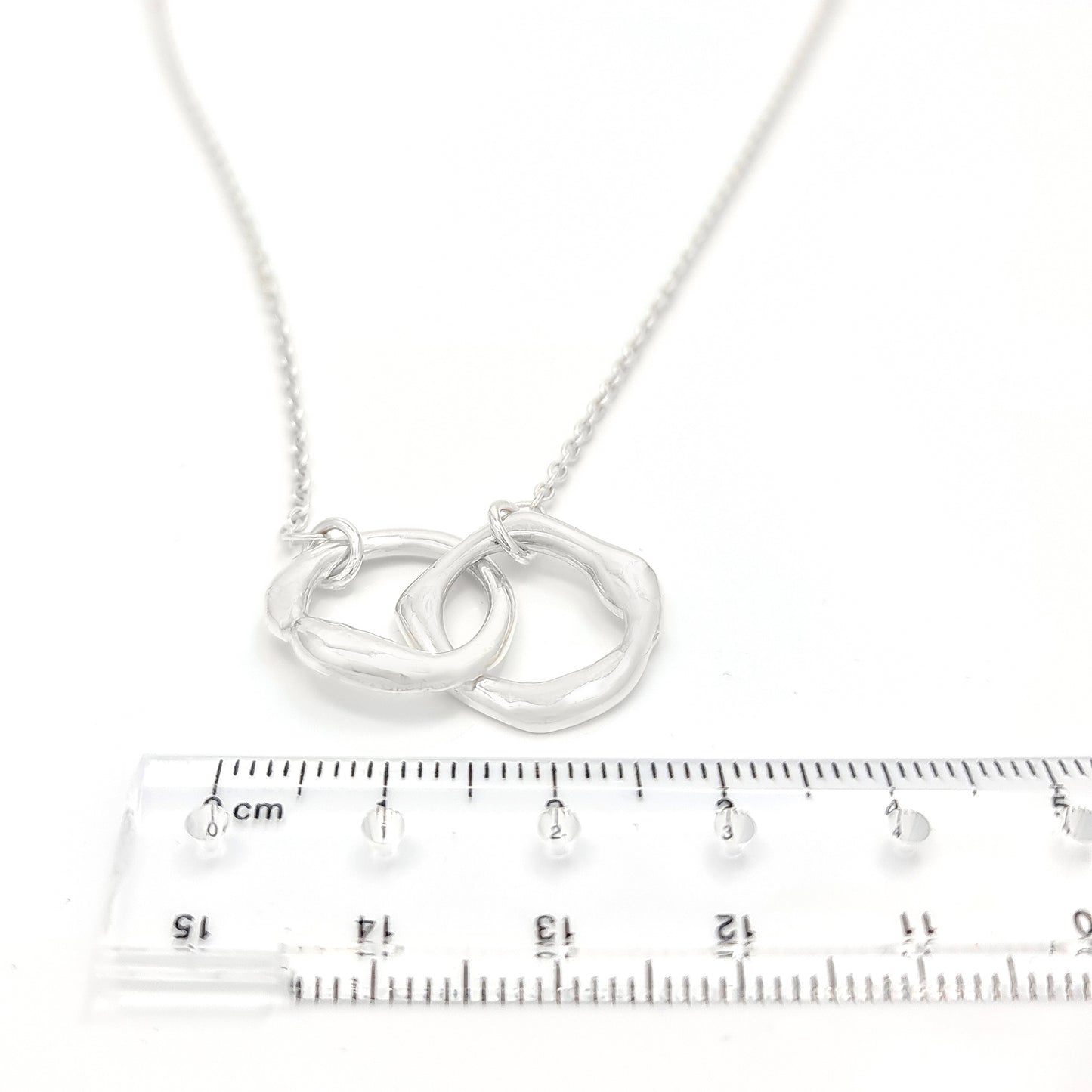 The Hoop Necklace by FR | Art Jewellery is a silver piece with a delicate chain that features two interlocking circular pendants. The pendants have a slightly irregular, textured appearance, creating a modern yet organic look. This handmade jewellery piece is laid flat against a plain white background.