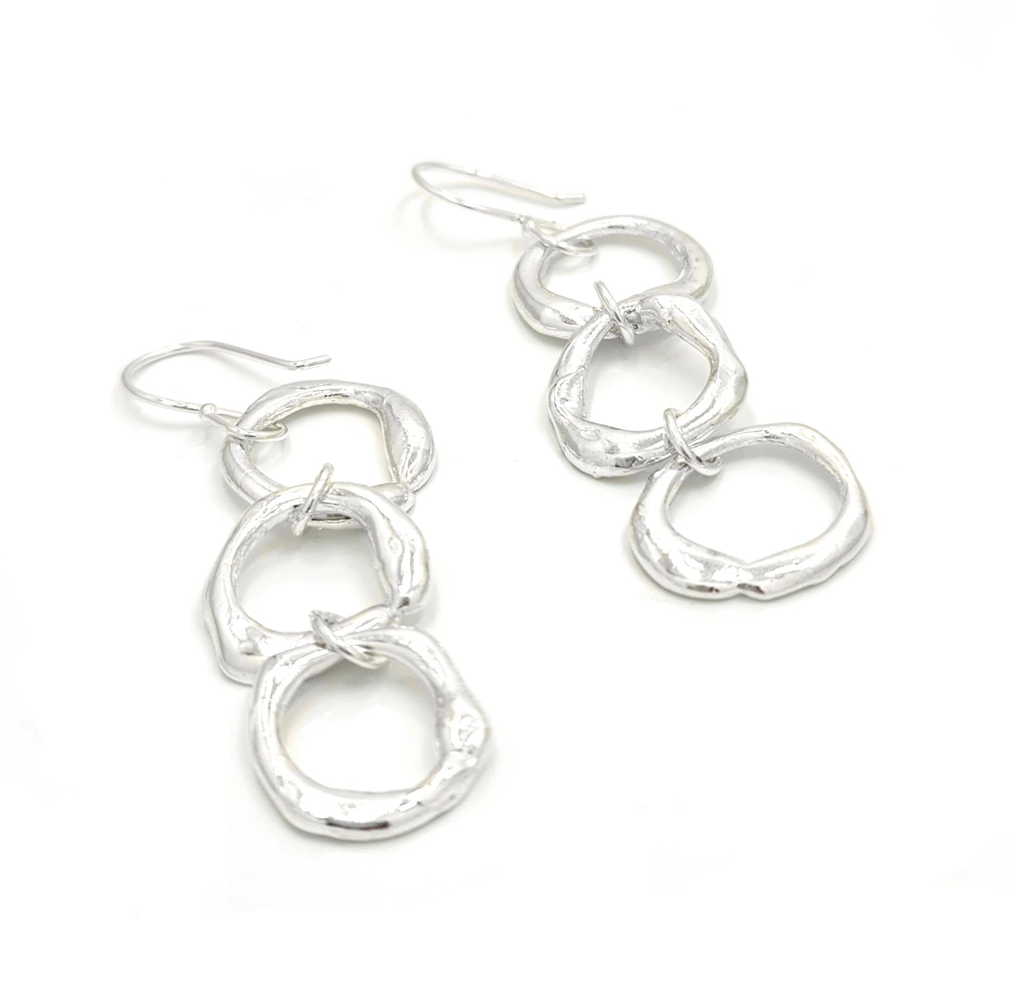 Introducing the "Join Together Earrings" by FR | Art Jewellery: a pair of elegant dangle earrings featuring three interlocking, irregularly shaped silver rings. Handmade with precision, these rings are linked vertically to create a fluid and contemporary design. The earrings also come with simple hook fastenings at the top for easy wear.