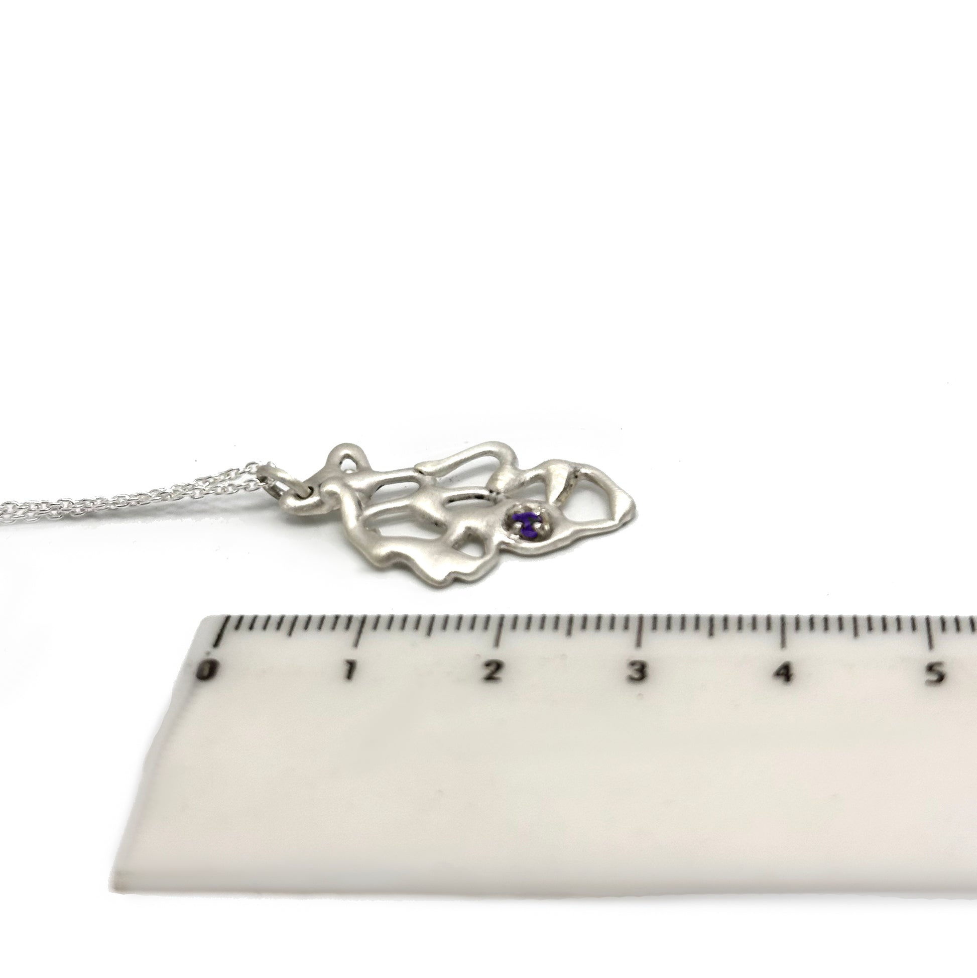 The Leaf Necklace by FR | Art Jewellery is displayed next to a ruler with centimeter markings, showcasing its abstract design and small purple gemstone. The sterling silver chain of the necklace is partially visible, and the white background enhances the intricate details of the pendant.