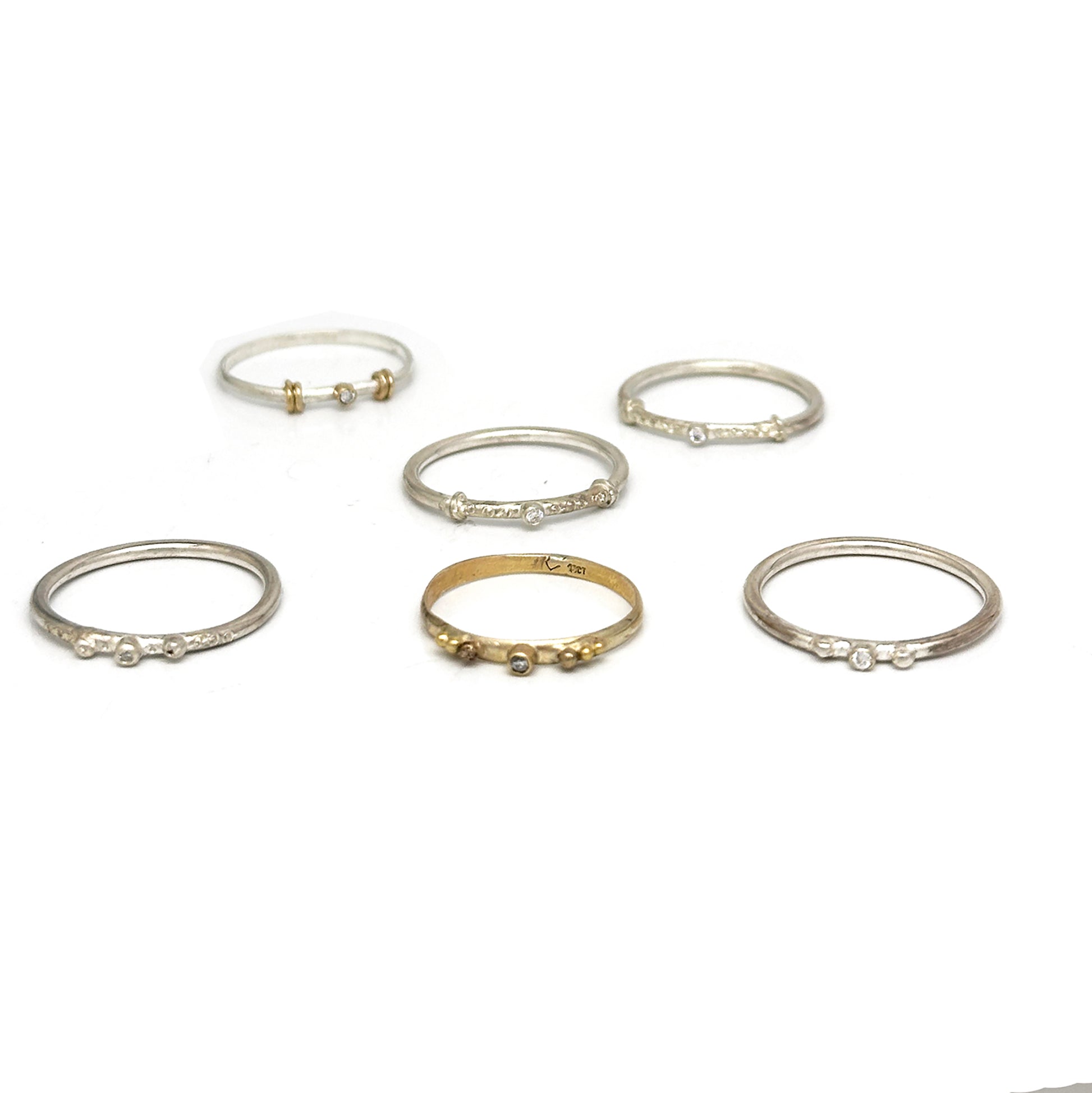 A collection of six Miniature Rings from FR | Art Jewellery is displayed on a white background. Five of the rings are crafted from silver with small gem accents, and one is gold, featuring a slightly larger central stone. Each handmade piece exhibits its own unique design and arrangement of stones.