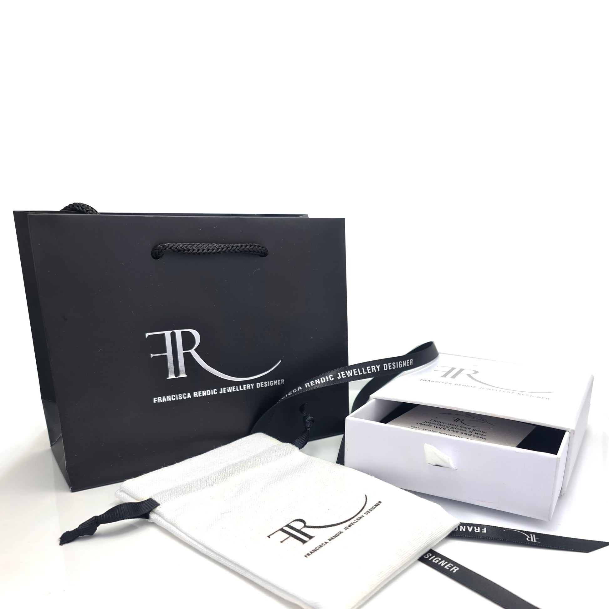 A selection of packaging items from FR | Art Jewellery Gift Wrapping is displayed. Included are a black shopping bag with handles, a white drawstring pouch, and a partially open white box revealing its contents. Each item prominently features the designer's logo, offering an elegant presentation ideal for a memorable unwrapping experience.