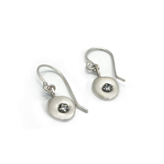 Introducing the Seed Earrings by FR | Art Jewellery: a pair of sterling silver hook earrings featuring gracefully suspended, round, flat discs. Each disc is elegantly adorned with a small, glittering gemstone at its center. As part of our Blossom Series, these earrings showcase a modern and minimalist design, artfully presented against a simple white background.