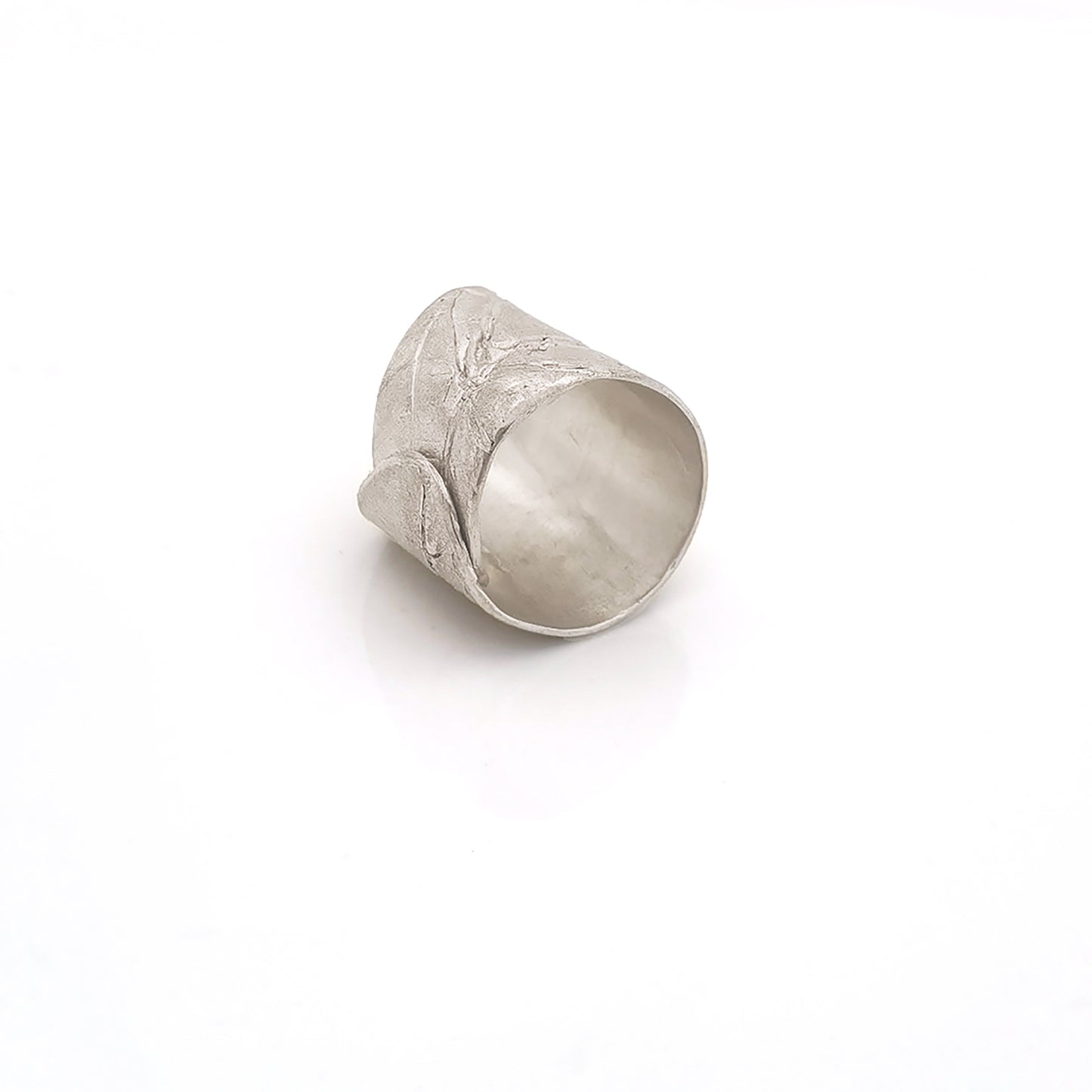 The FR | Art Jewellery Silver Leaf Ring is a wide, textured silver band with a matte finish. Its organic, slightly irregular shape and subtle line impressions contribute to its rustic aesthetic. As a piece of handmade jewellery, the ring glistens slightly due to the light reflecting off its uneven surface.