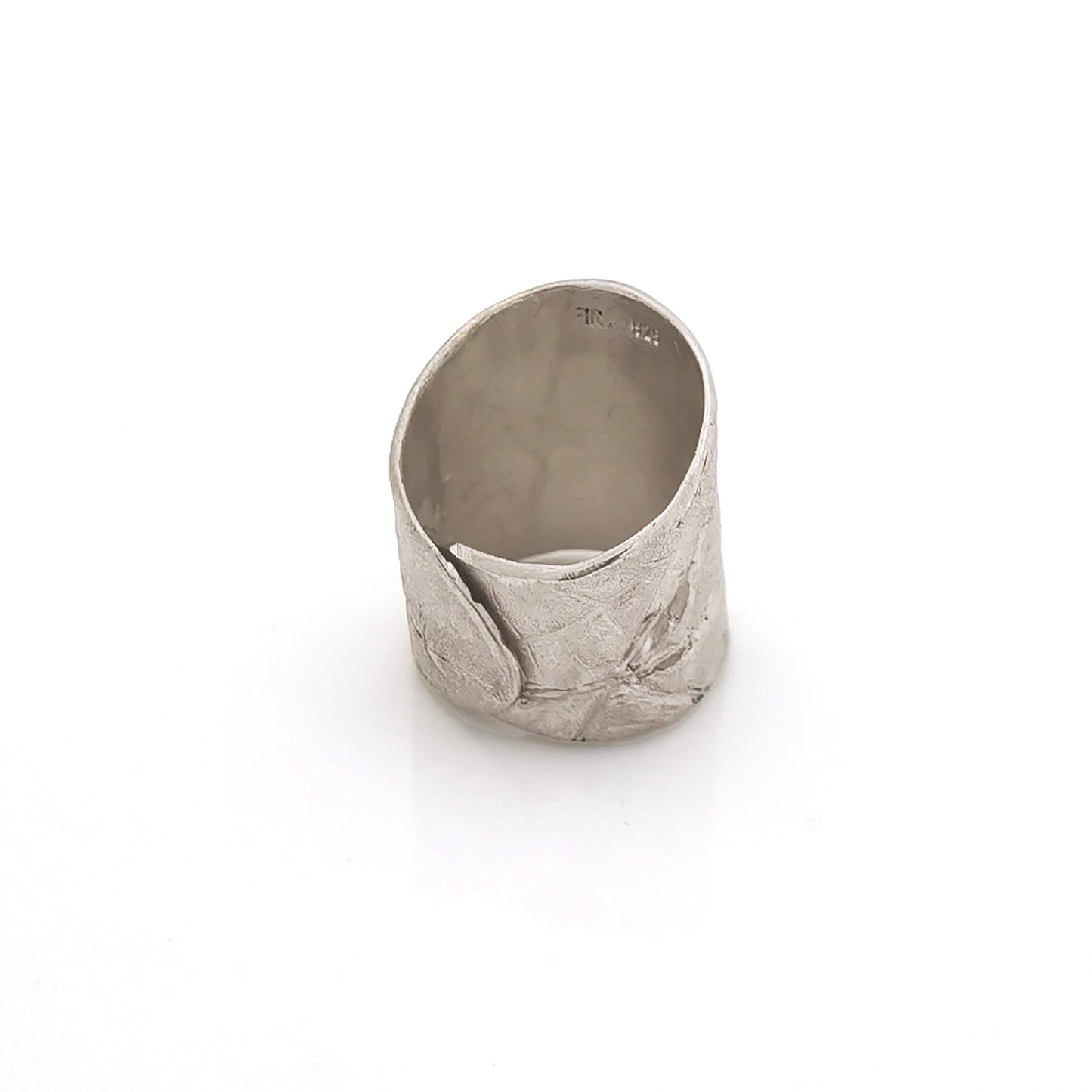 The FR | Art Jewellery Silver Leaf Ring is a wide, textured silver band with a matte finish. Its organic, slightly irregular shape and subtle line impressions contribute to its rustic aesthetic. As a piece of handmade jewellery, the ring glistens slightly due to the light reflecting off its uneven surface.