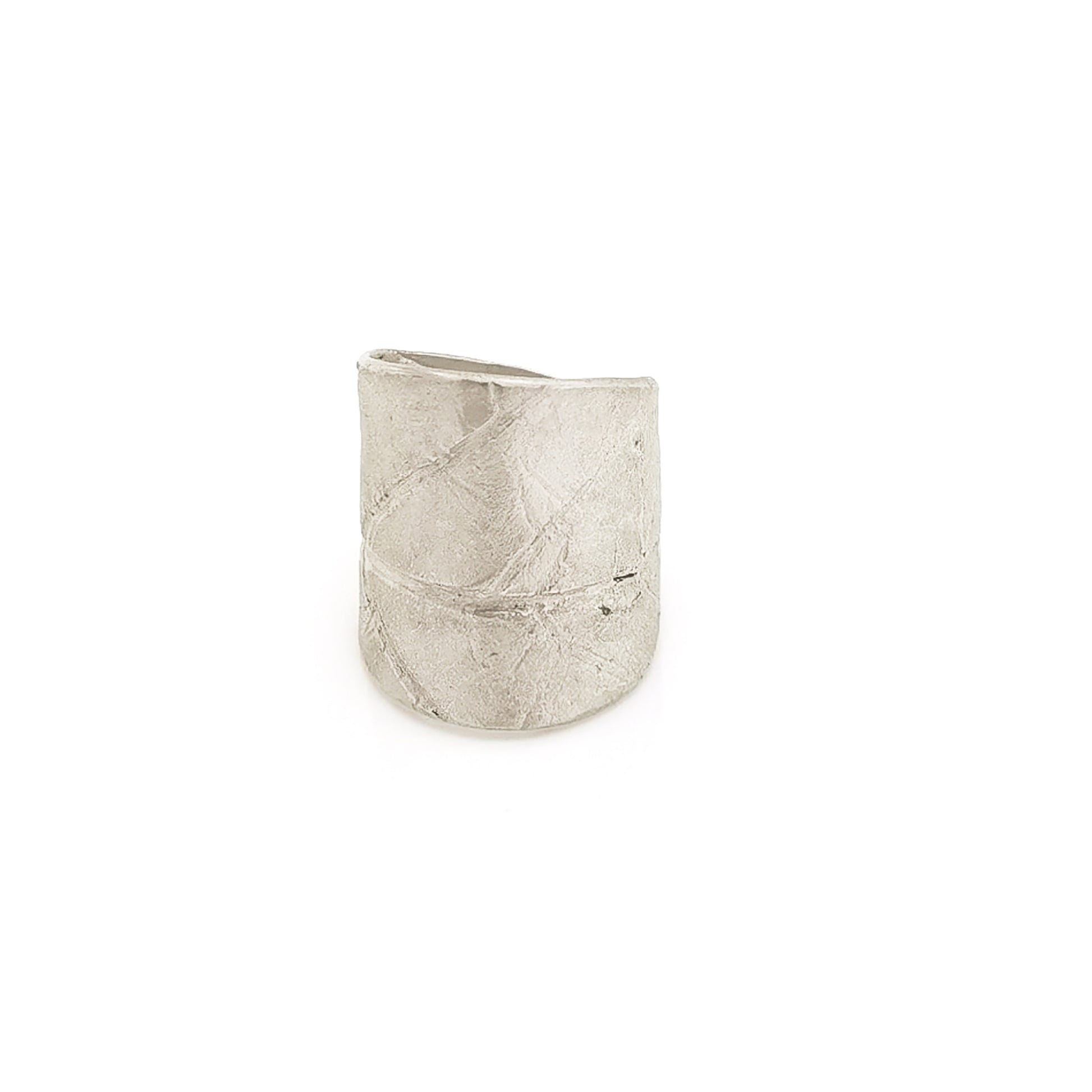 The FR | Art Jewellery Silver Leaf Ring is a wide, textured silver band with a matte finish. Its organic, slightly irregular shape and subtle line impressions contribute to its rustic aesthetic. As a piece of handmade jewellery, the ring glistens slightly due to the light reflecting off its uneven surface.