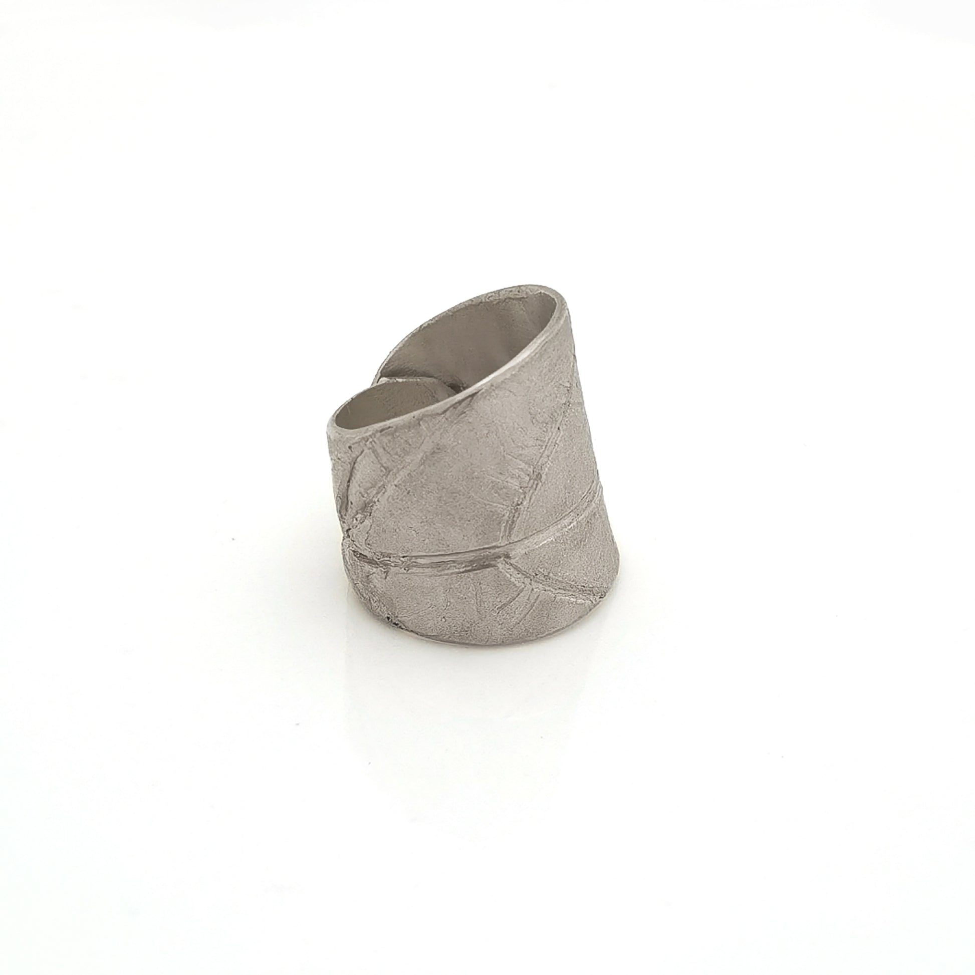 The FR | Art Jewellery Silver Leaf Ring is a wide, textured silver band with a matte finish. Its organic, slightly irregular shape and subtle line impressions contribute to its rustic aesthetic. As a piece of handmade jewellery, the ring glistens slightly due to the light reflecting off its uneven surface.