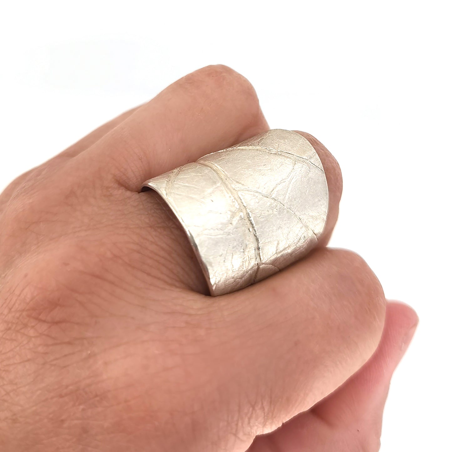 The FR | Art Jewellery Silver Leaf Ring is a wide, textured silver band with a matte finish. Its organic, slightly irregular shape and subtle line impressions contribute to its rustic aesthetic. As a piece of handmade jewellery, the ring glistens slightly due to the light reflecting off its uneven surface.