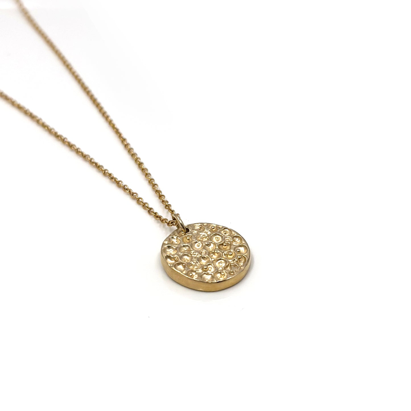 The Spot Necklace Big from FR | Art Jewellery is a silver necklace featuring a delicate chain and a round pendant. The pendant has a textured surface adorned with small, raised circular shapes that create an intricate pattern. Coordinating handmade jewellery, including custom-made gold-plated earrings, is also available to complement this exquisite piece.