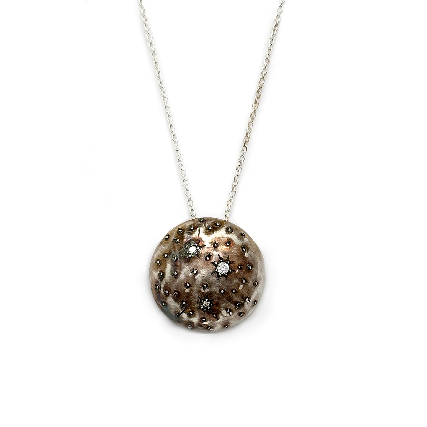 The Starburst Necklace by FR | Art Jewellery features a round, textured pendant on a thin silver chain. The pendant boasts a hammered finish with several small, sparkling accents embedded within its surface, mimicking a celestial or cratered appearance. This piece exemplifies the artistry found in handmade jewelry.