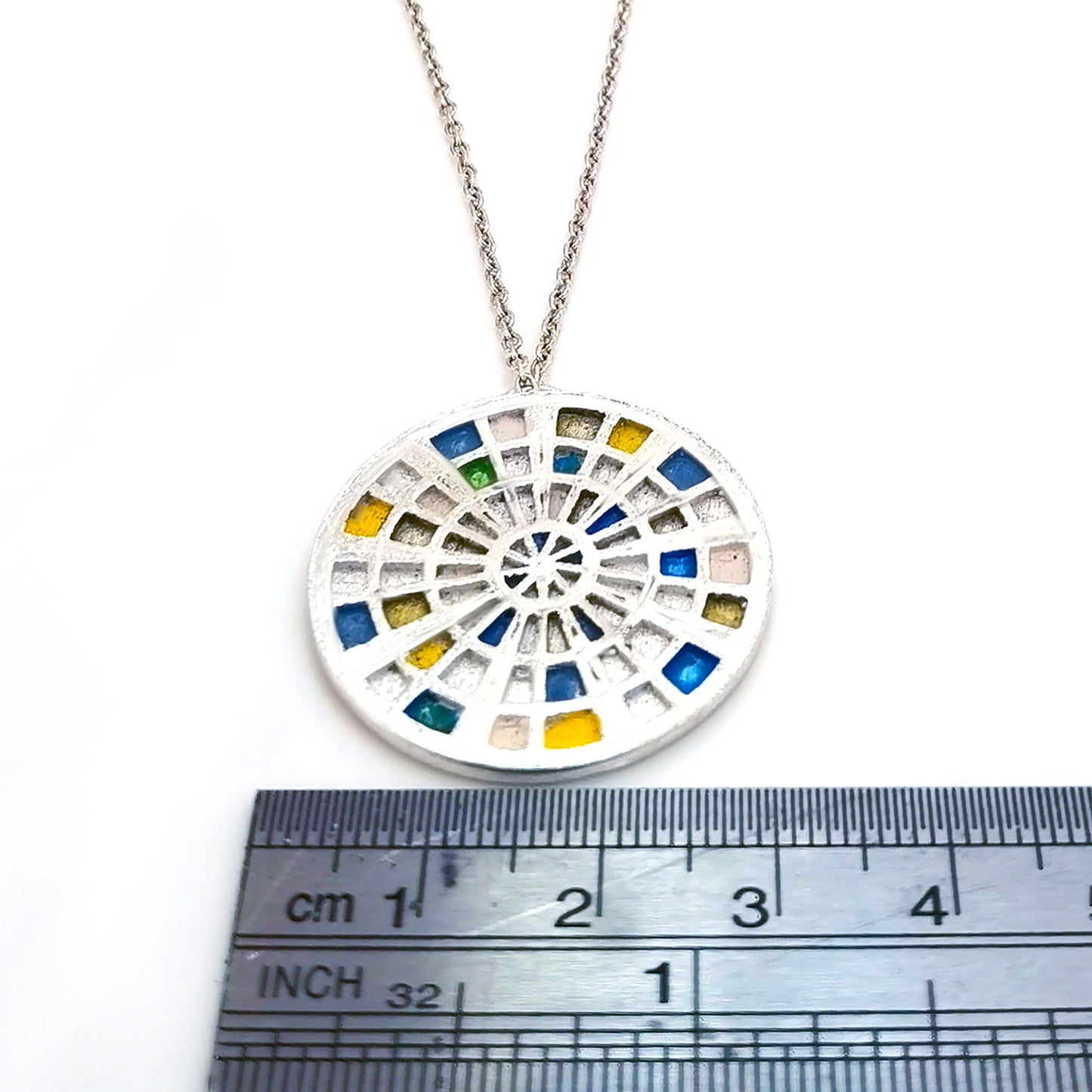 Introducing the "Talis Necklace blue 70cms" by FR | Art Jewellery—an elegant silver piece adorned with a round pendant that features an intricate stained glass window design. This mosaic pattern is comprised of small blue, green, yellow, and white squares arranged in a captivating circular layout. For the perfect ensemble, pair it with our gold-plated earrings or explore our custom-made options for unique handmade jewellery pieces.