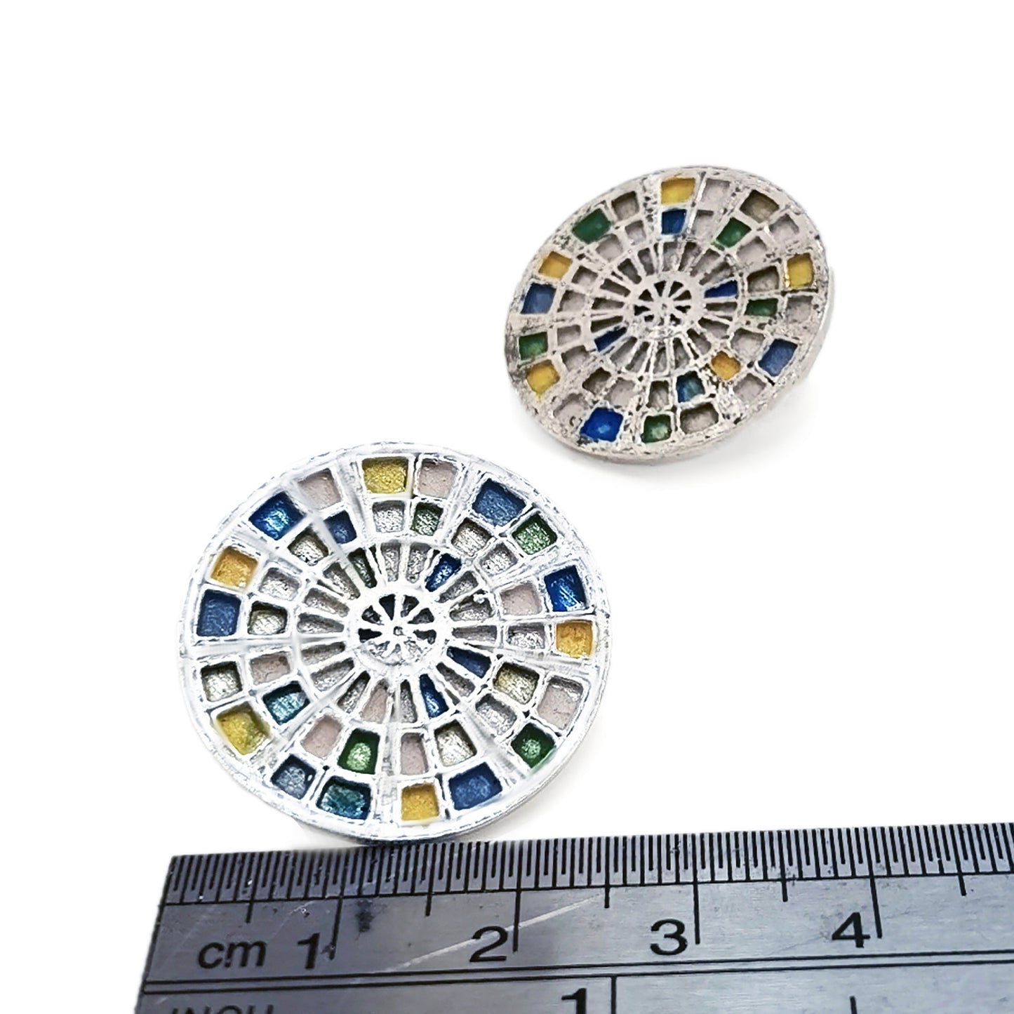 Introducing the Talis Earrings by FR | Art Jewellery, a stunning pair of round stud earrings designed with a stained glass window motif. These intricate pieces feature colored segments in shades of blue, green, and yellow, all beautifully set in silver-tone metal. Handmade to perfection, one earring is displayed front-on while the other is subtly angled.