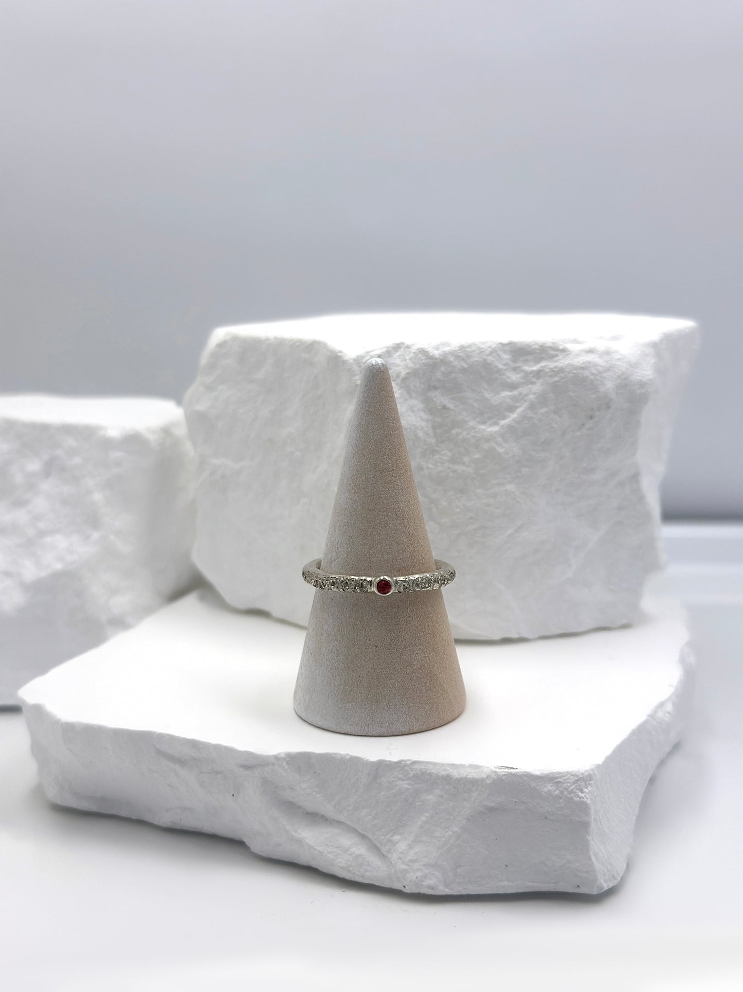 The FR | Art Jewellery Tinytoo Sterling Silver Ring with Ruby Gemstone features a textured band and a small round ruby, elegantly displayed on a white background.