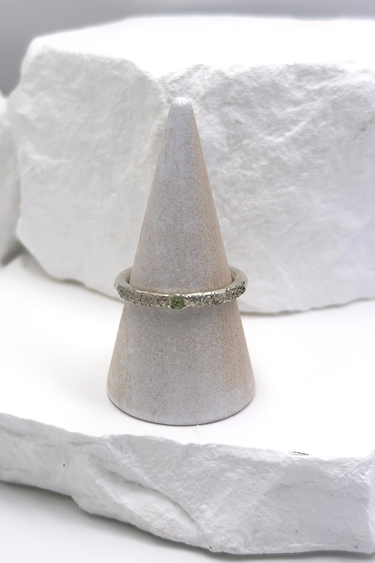 The FR | Art Jewellery Tinytoo Sterling Silver Ring with Peridot Gemstone features a green stone and textured band, shown on a white cone stand against a white stone background.