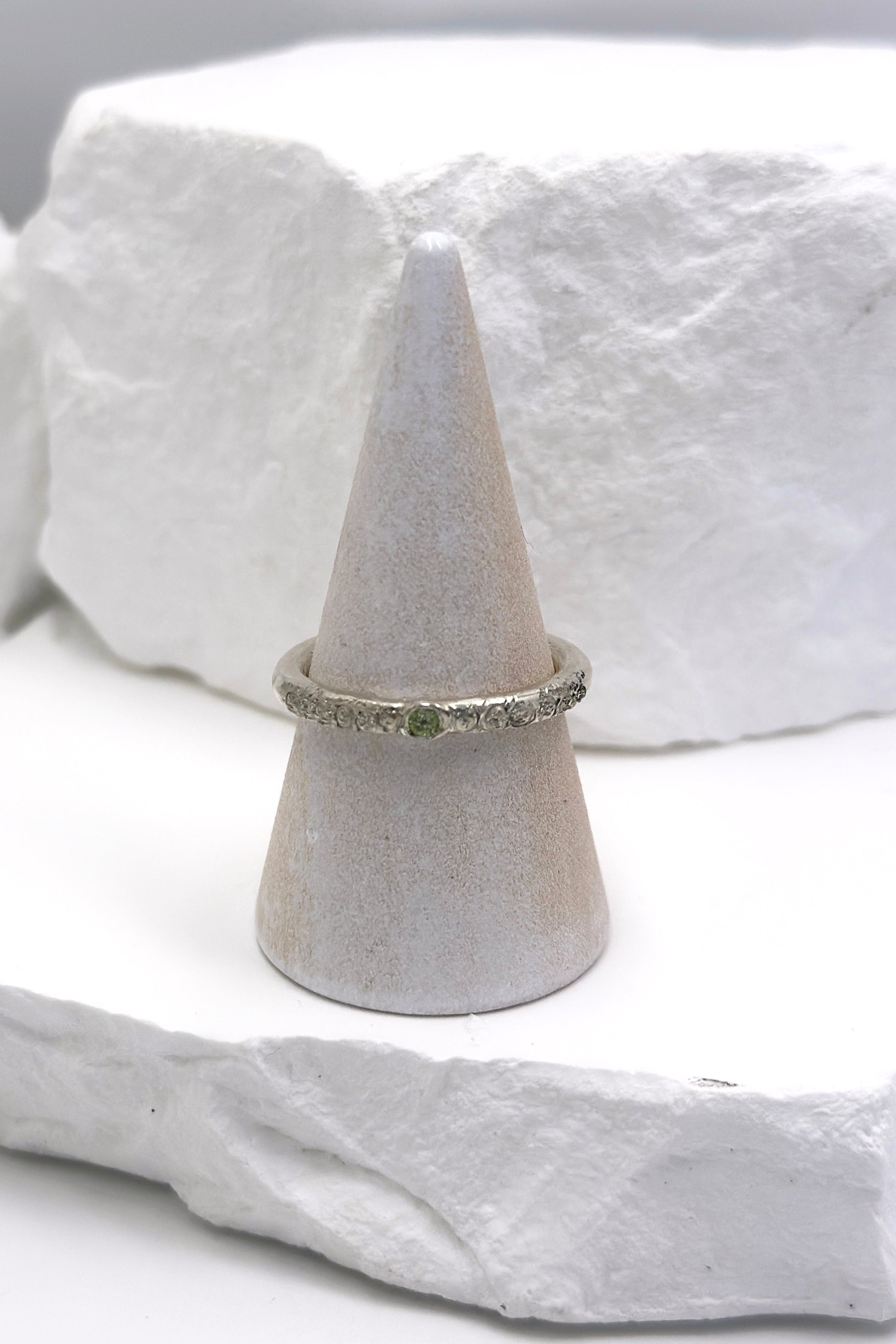 The FR | Art Jewellery Tinytoo Sterling Silver Ring with Peridot Gemstone features a green stone and textured band, shown on a white cone stand against a white stone background.