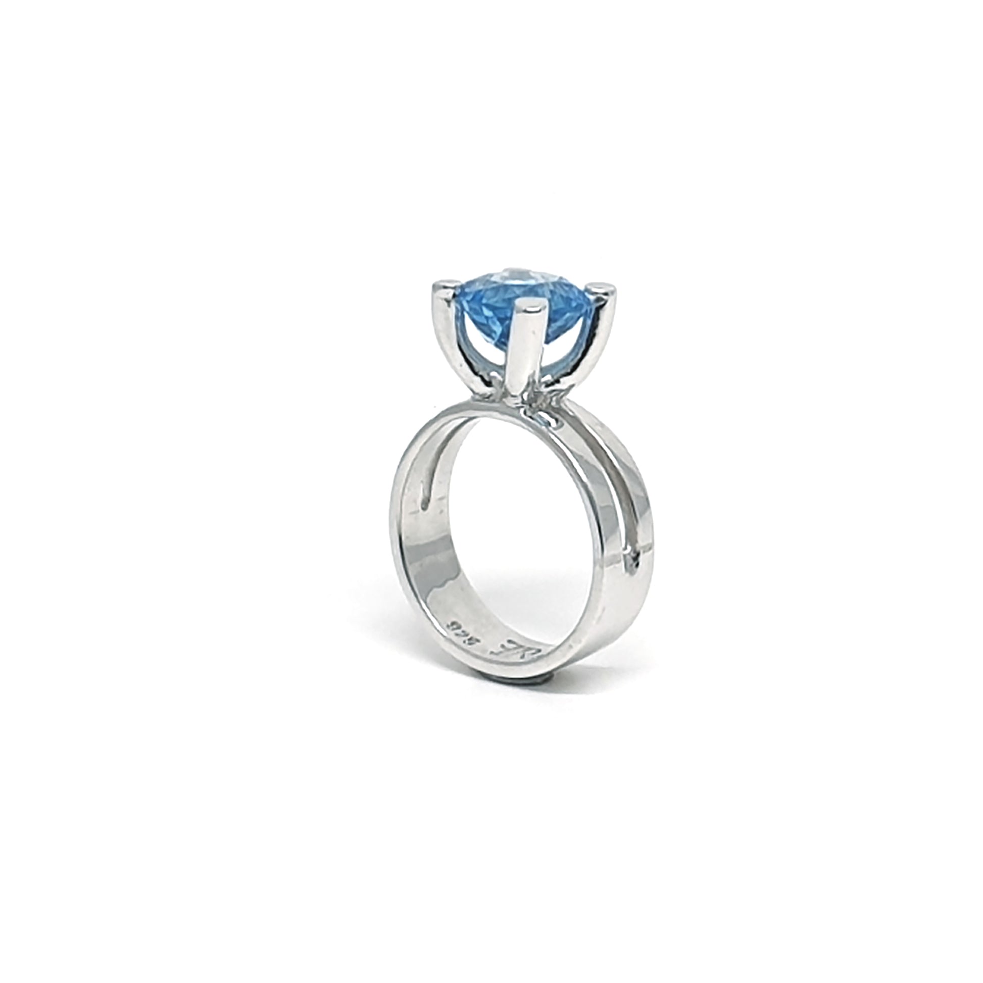 The Topaz Ring Claw from FR | Art Jewellery features a stunning, large round blue topaz gemstone set in a simple, elegant silver design. The topaz is prominently displayed in a four-prong setting, elevating it above the ring band and showcasing the exceptional craftsmanship of our handmade jewellery collection. The accompanying image displays the piece against a white background.