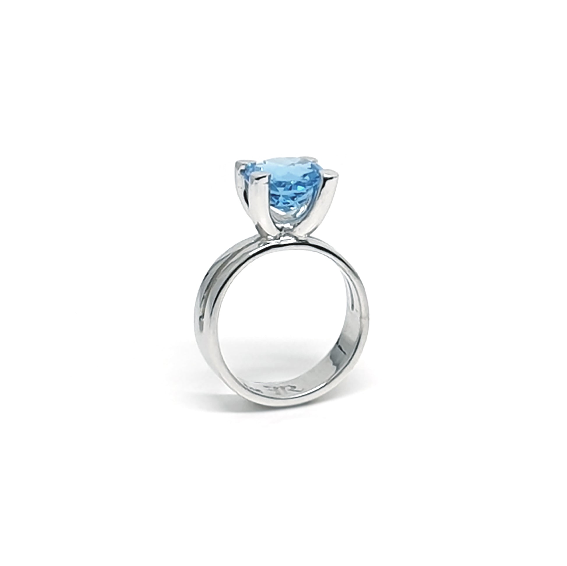 The Topaz Ring Claw from FR | Art Jewellery features a stunning, large round blue topaz gemstone set in a simple, elegant silver design. The topaz is prominently displayed in a four-prong setting, elevating it above the ring band and showcasing the exceptional craftsmanship of our handmade jewellery collection. The accompanying image displays the piece against a white background.