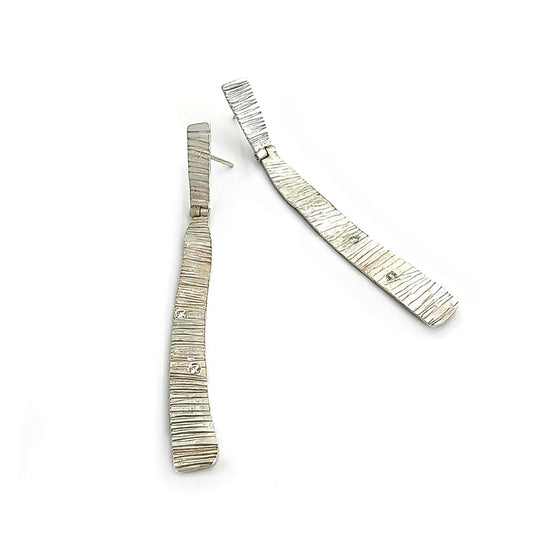 The Tribu Diamonds Earrings by FR | Art Jewellery are elongated, rectangular, sterling silver pieces featuring a textured, striped pattern. Each handmade earring is adorned with a small sparkling gemstone set within the design. These contemporary and artistic earrings have a slight curve, creating a dynamic and elegant look.